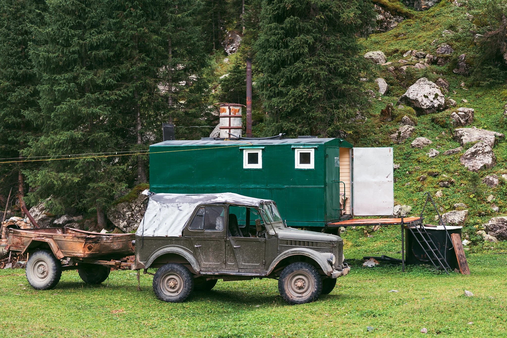 Old, rusty truck with a covered bed parked in front of a green container on a grassy area with trees and rocks in the background.