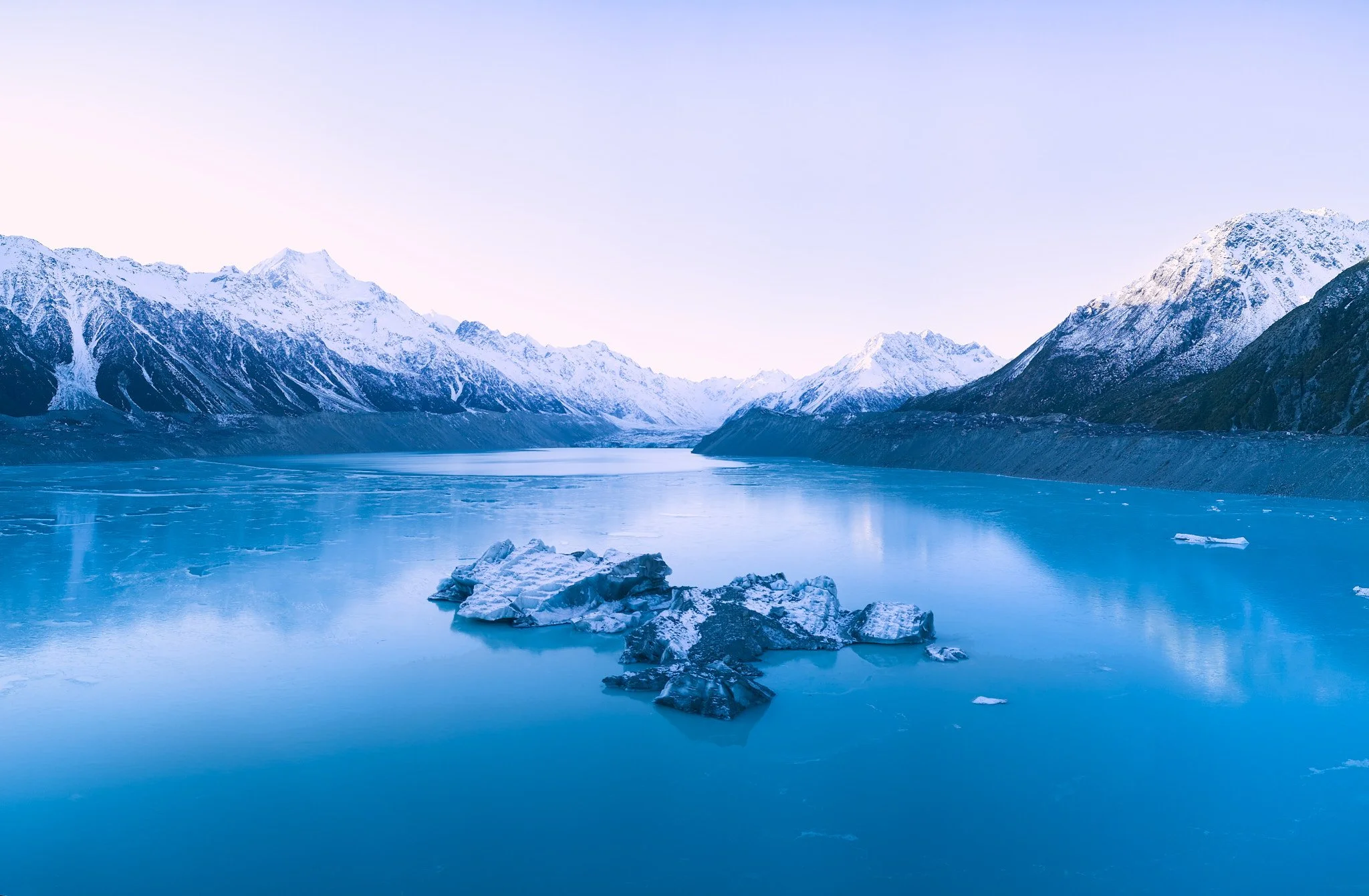 Snow-capped mountains surrounding a calm, icy blue glacier lake with floating ice chunks.