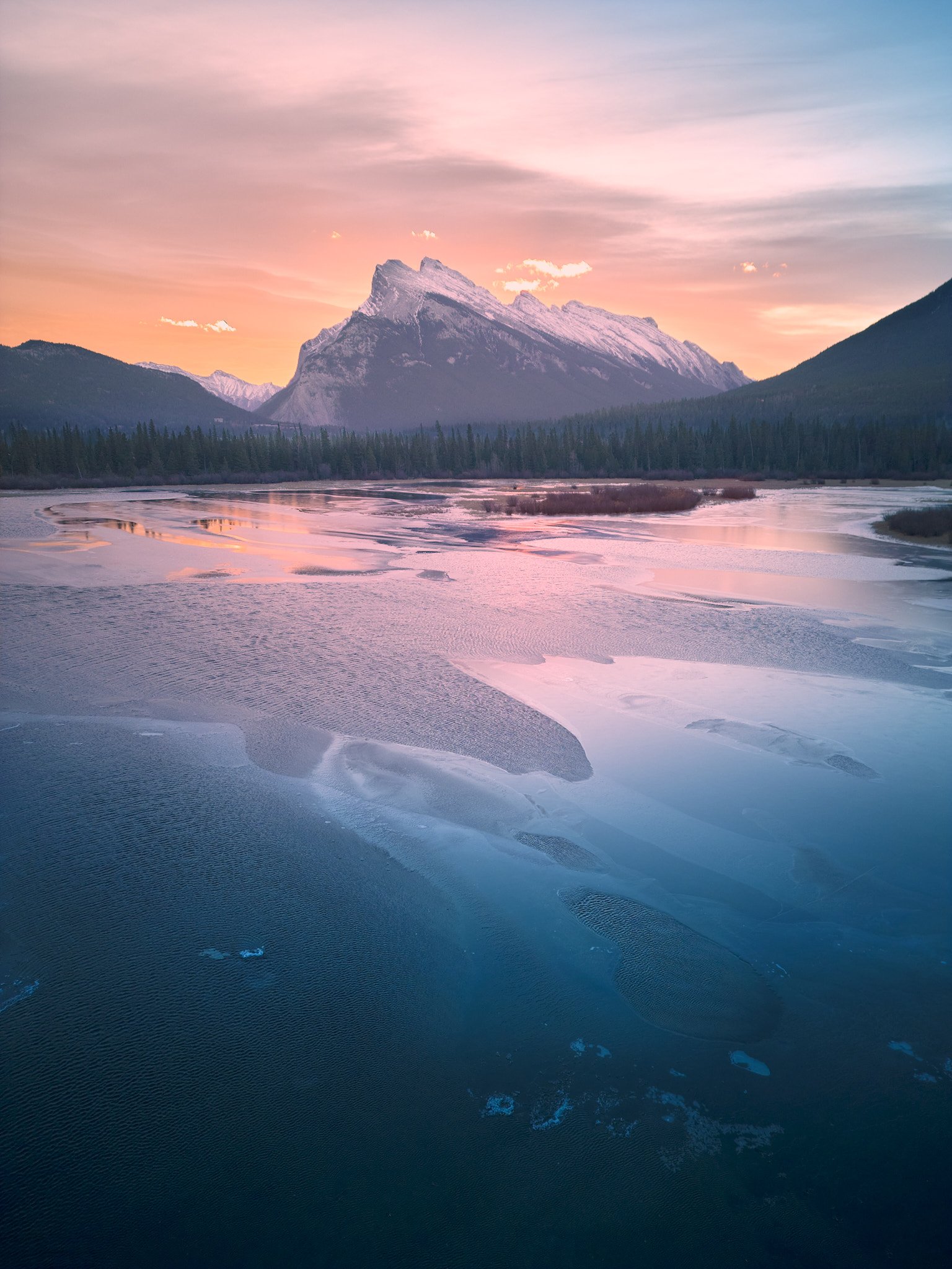 A mountain landscape at sunset with snow-capped peaks, a forest in the foreground, and a partially frozen river reflecting the pink and purple sky.