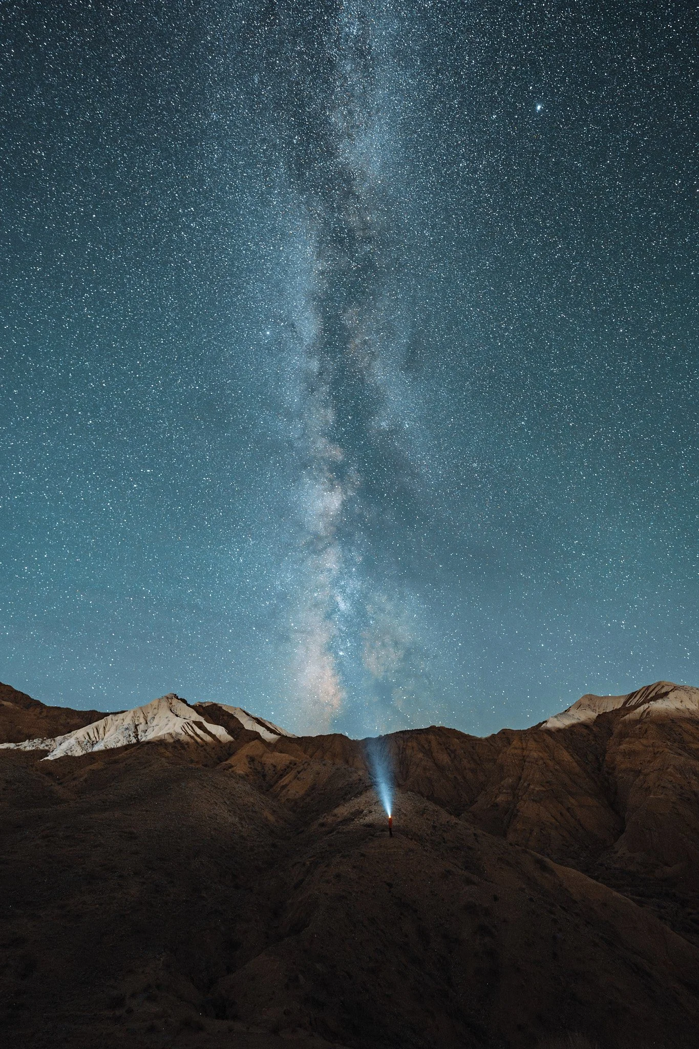 Night sky filled with stars and the Milky Way galaxy over mountainous terrain with a person holding a flashlight.