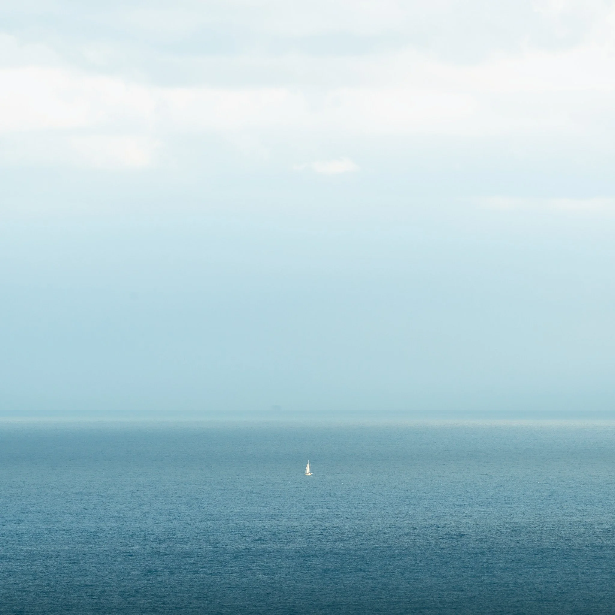 A vast ocean with a small sailboat in the distance under a cloudy sky.