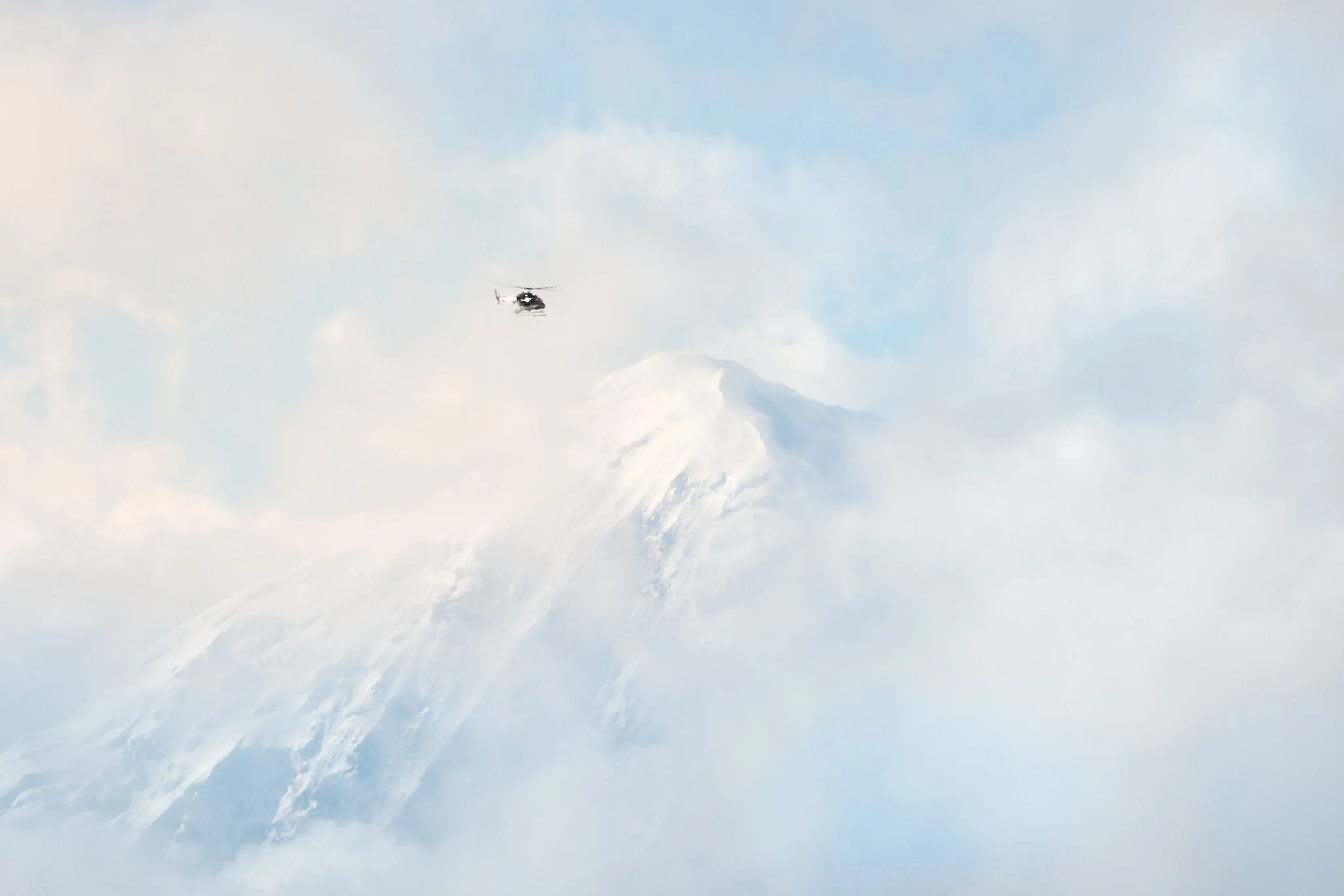 A helicopter flying near a snow-covered mountain peak, surrounded by clouds in Alaska
