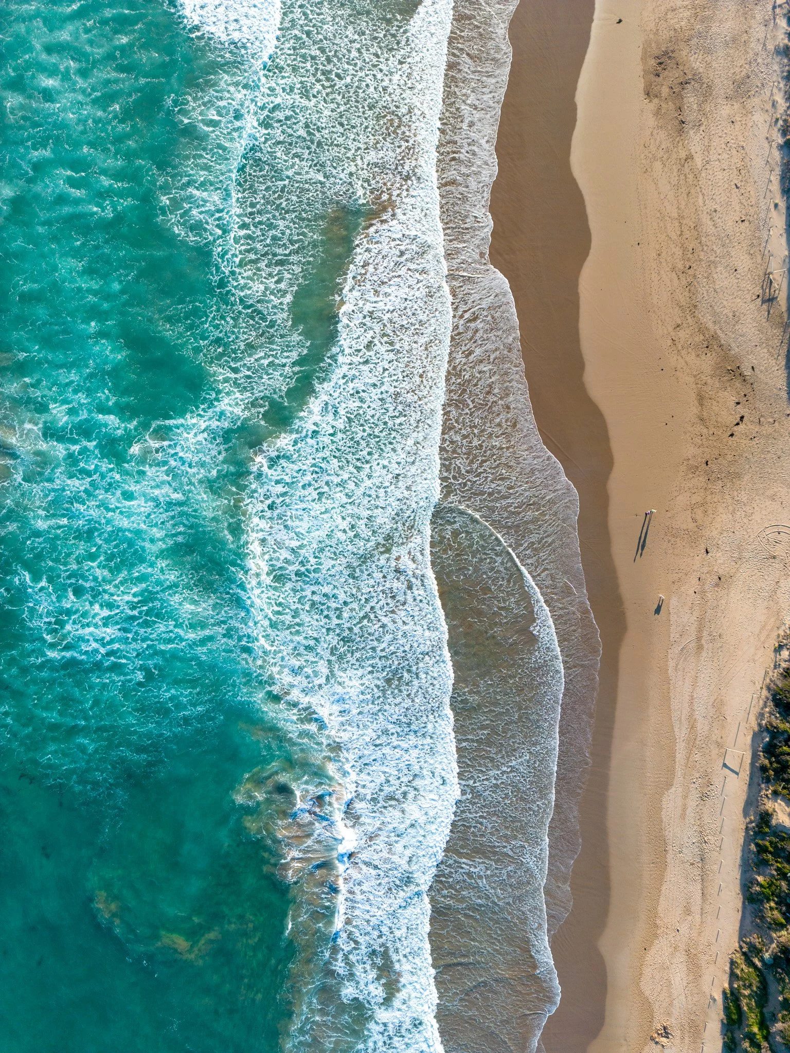 An aerial view of the shoreline at the beach with blue ocean waves crashing onto the sandy shore and surrounding greenery on the Great Ocean Road