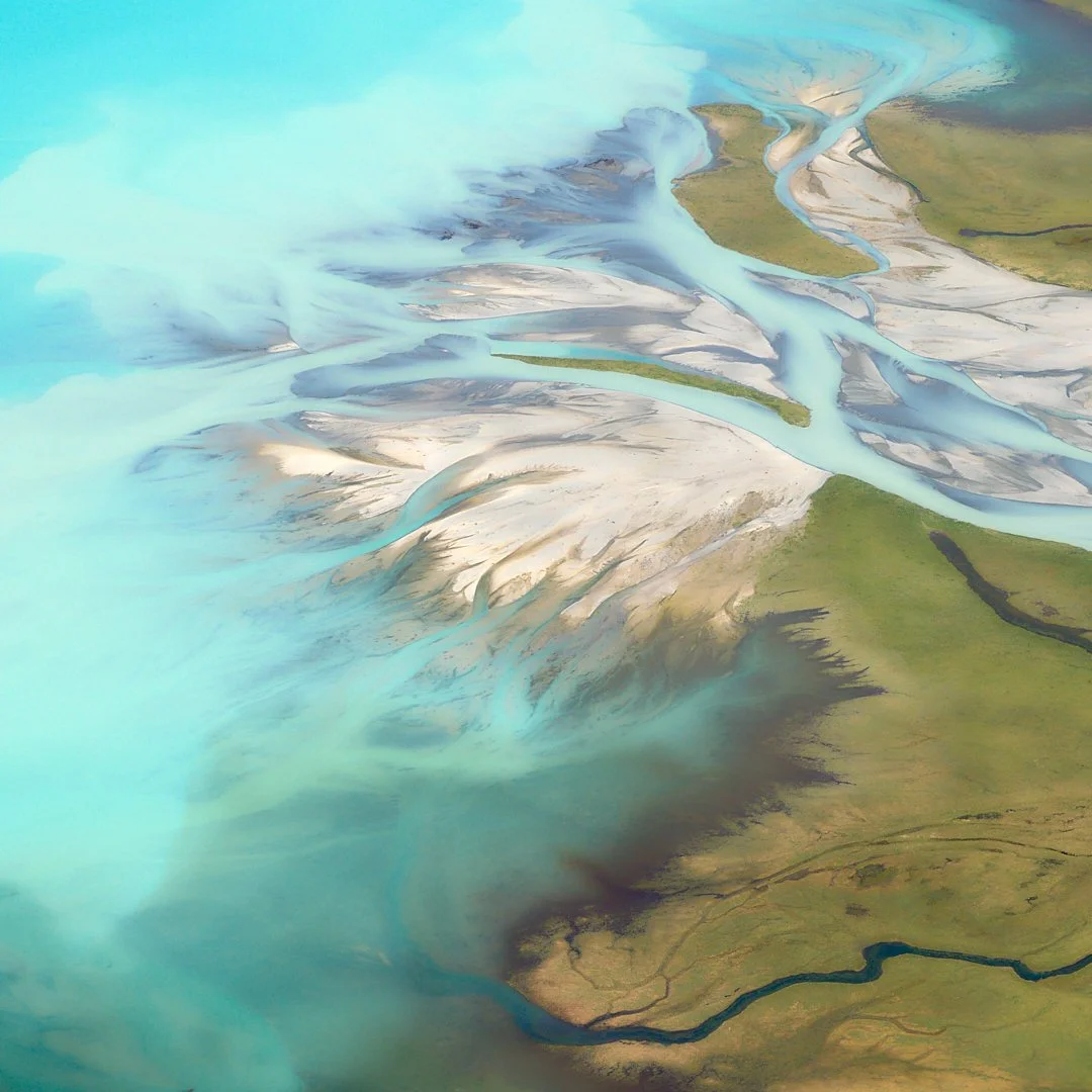 Aerial view of winding rivers and streams flowing through green and barren land, with sediment and glacial meltwater creating blue and white patterns.
