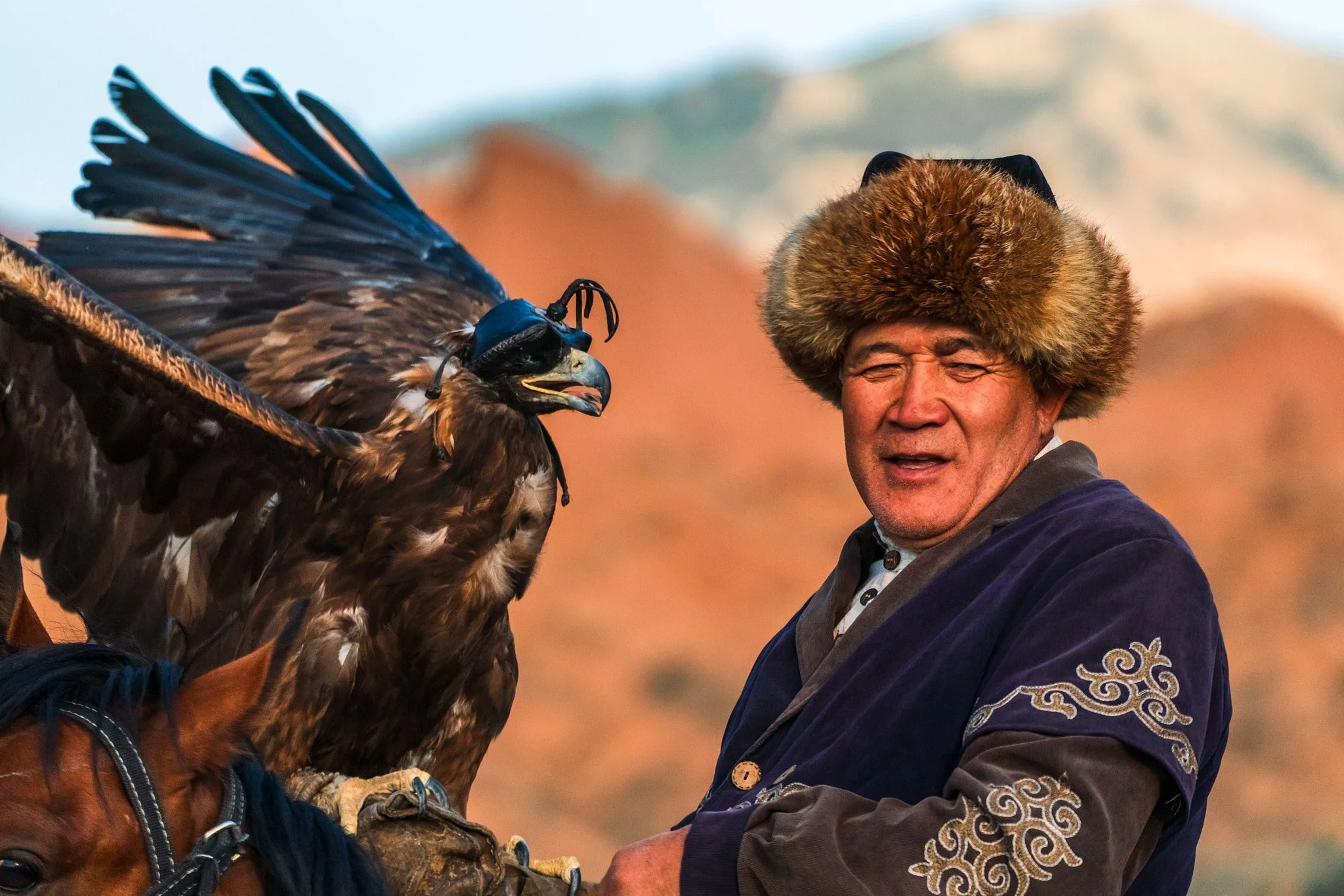 A man wearing a traditional fur hat and ornate clothing riding a horse with a large bird of prey perched on his arm, against a background of mountainous landscape.