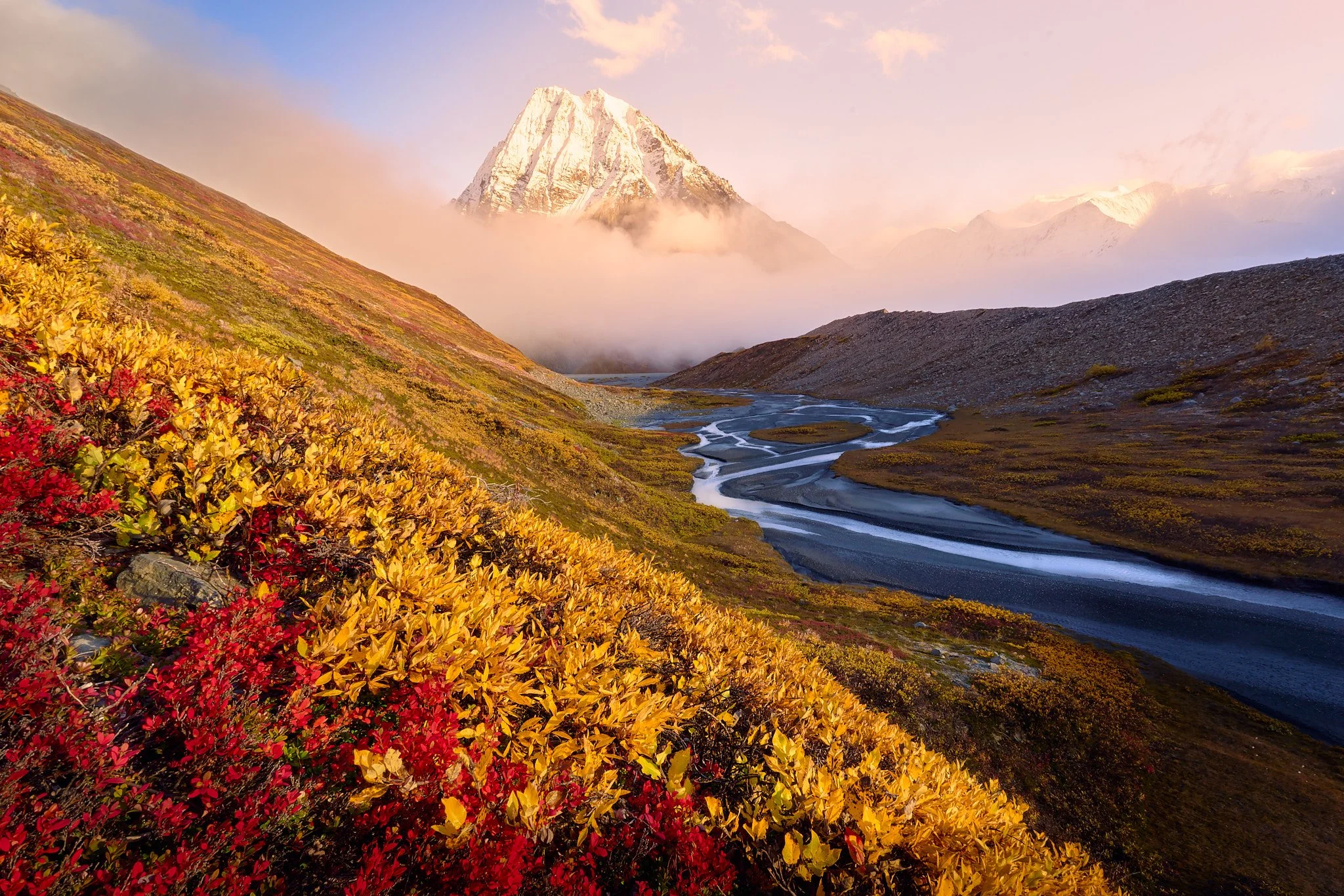A scenic mountain landscape with a snow-capped peak in the background, surrounded by clouds, and a winding river flowing through colorful autumn foliage in the foreground.
