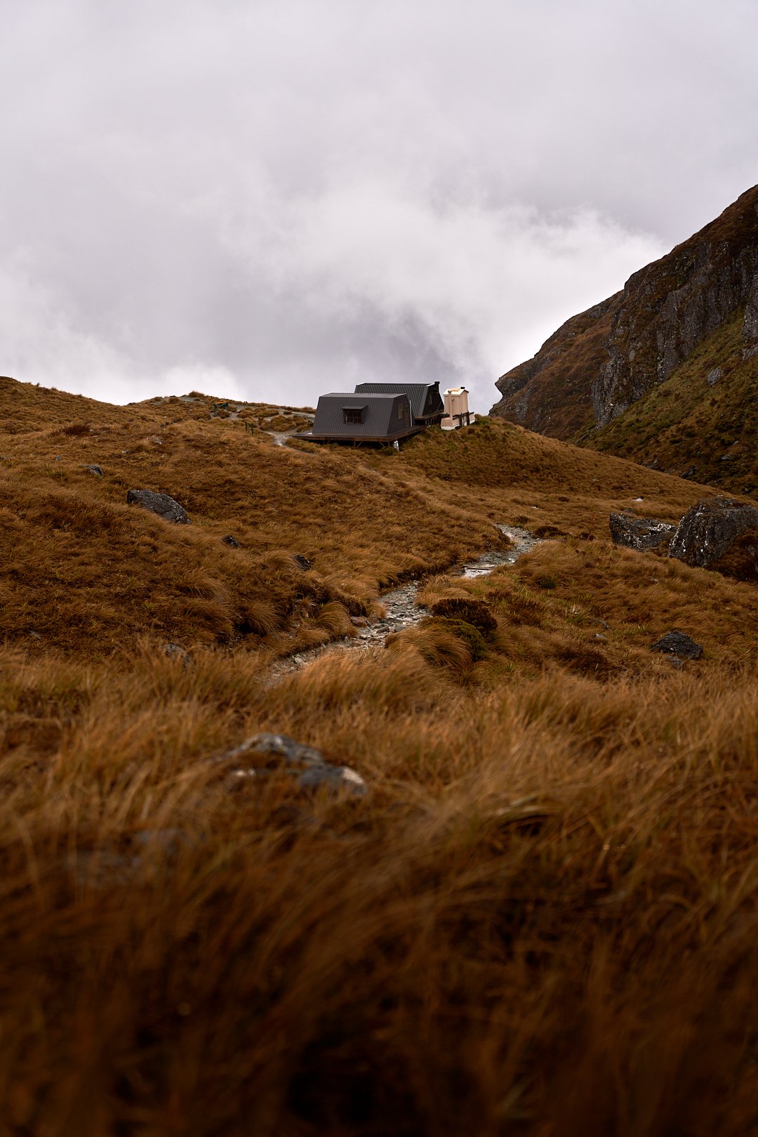 A secluded house on a hillside surrounded by tall, brown grass with a narrow stream running through the landscape and cloudy skies overhead.