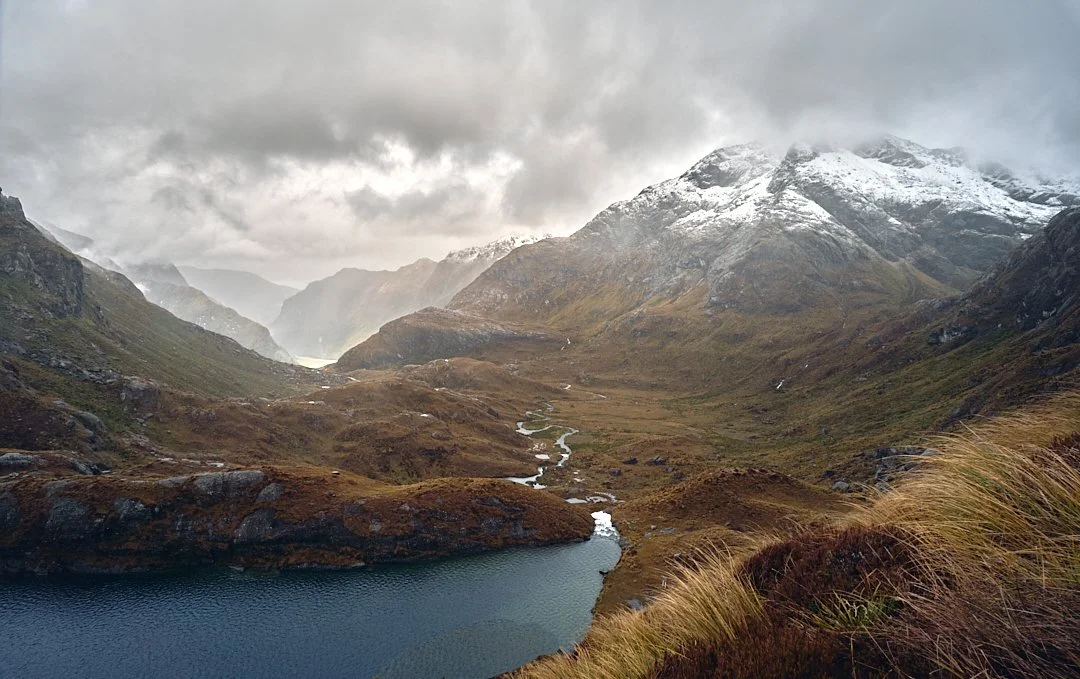 Mountain landscape with snow-capped peaks, a cloudy sky, a river flowing through the valley, and a lake in the foreground.