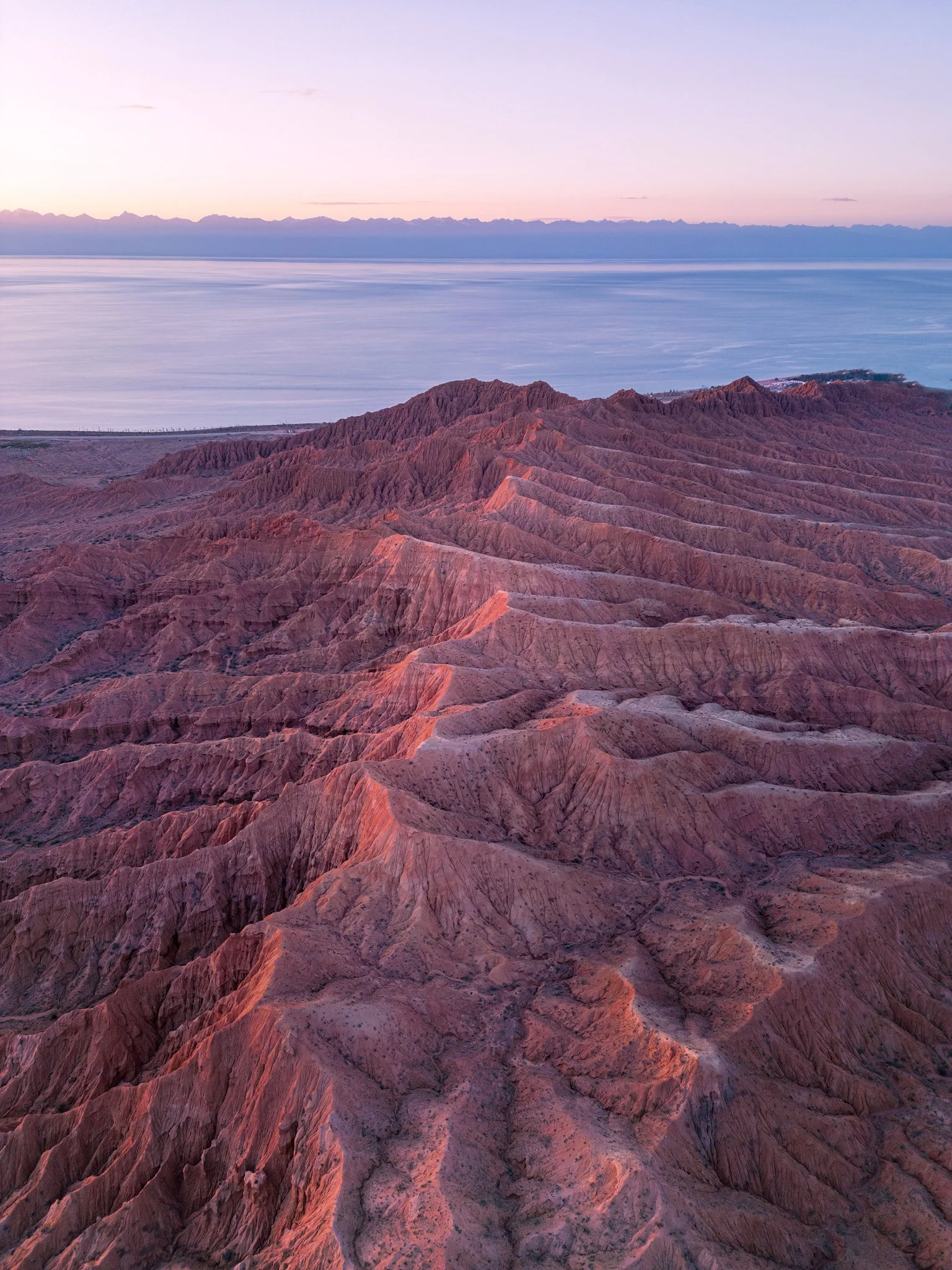 Colorful rocky mountains at sunset with a body of water and a distant mountain range in the background.