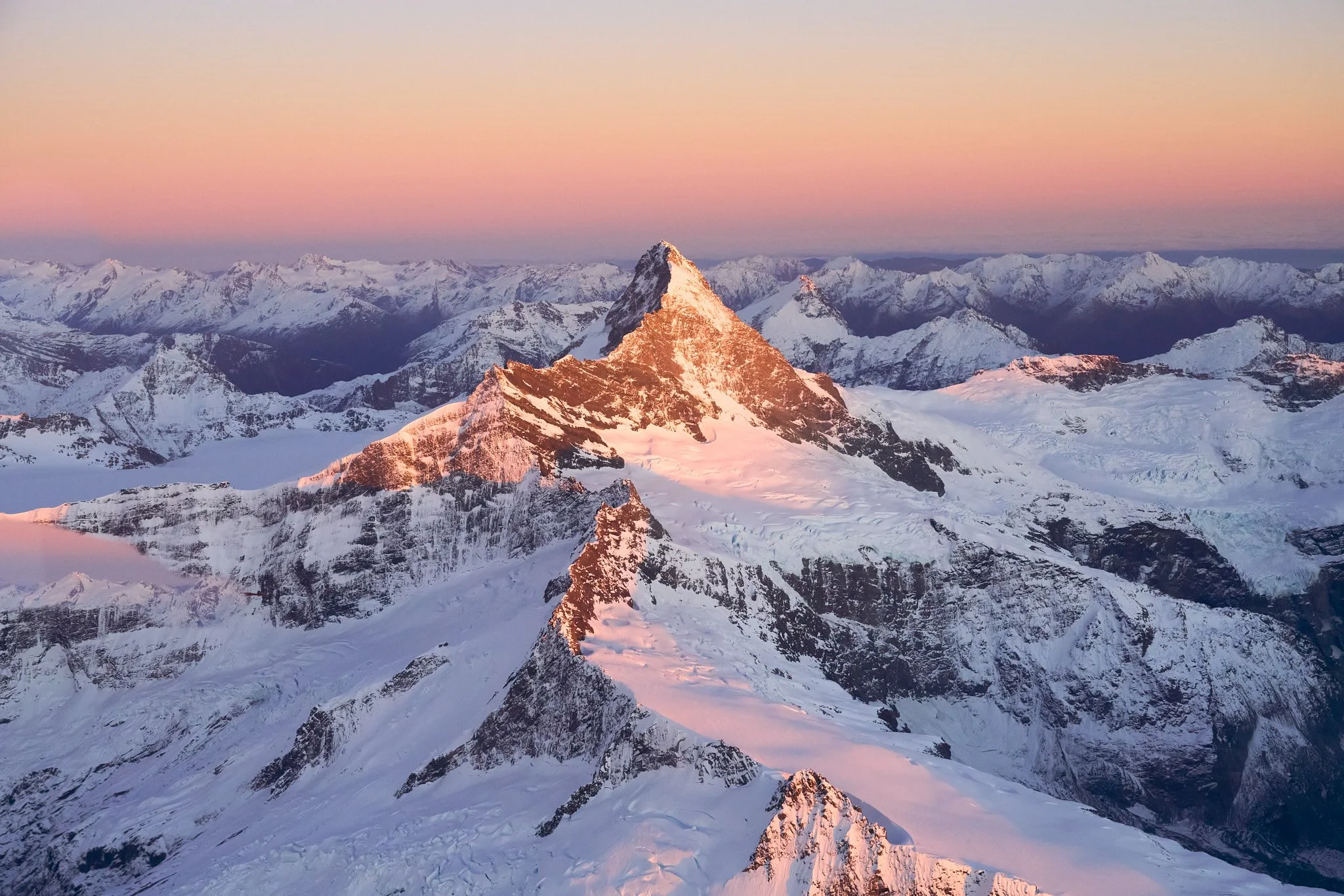 Snow-covered mountain peaks at sunset with pink and orange sky in the background.