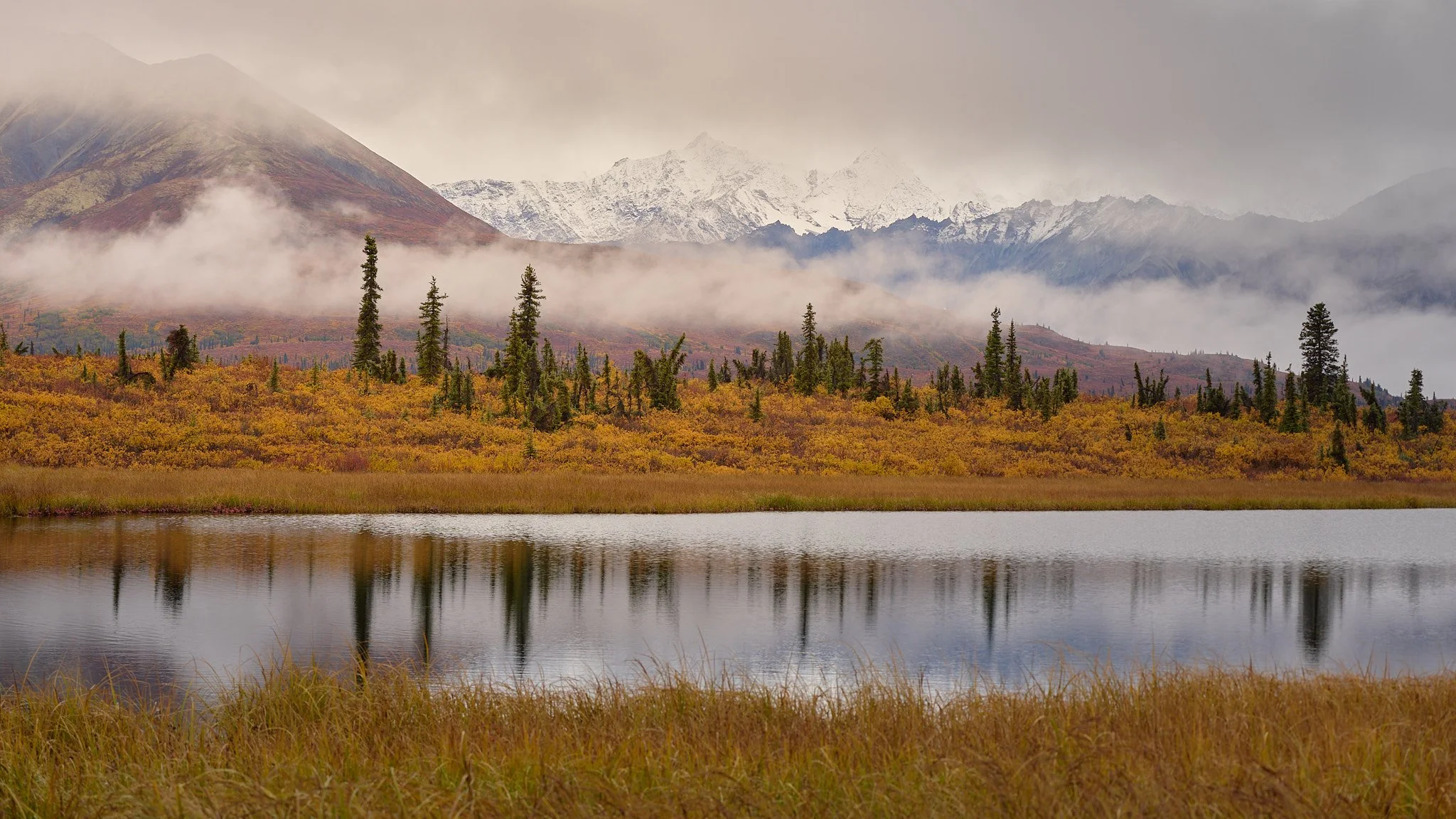 Mountain landscape with snow-capped peaks, clouds, and mist over a forest with autumn foliage and a calm lake in the foreground.