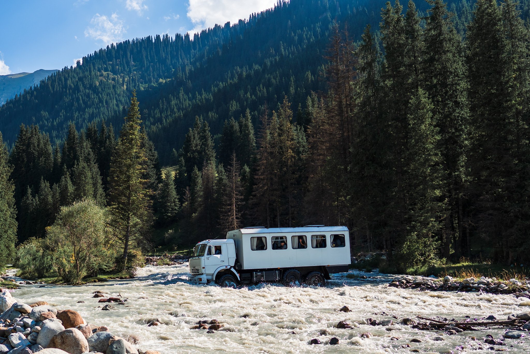 A white amphibious vehicle crossing a river surrounded by forest and mountains.