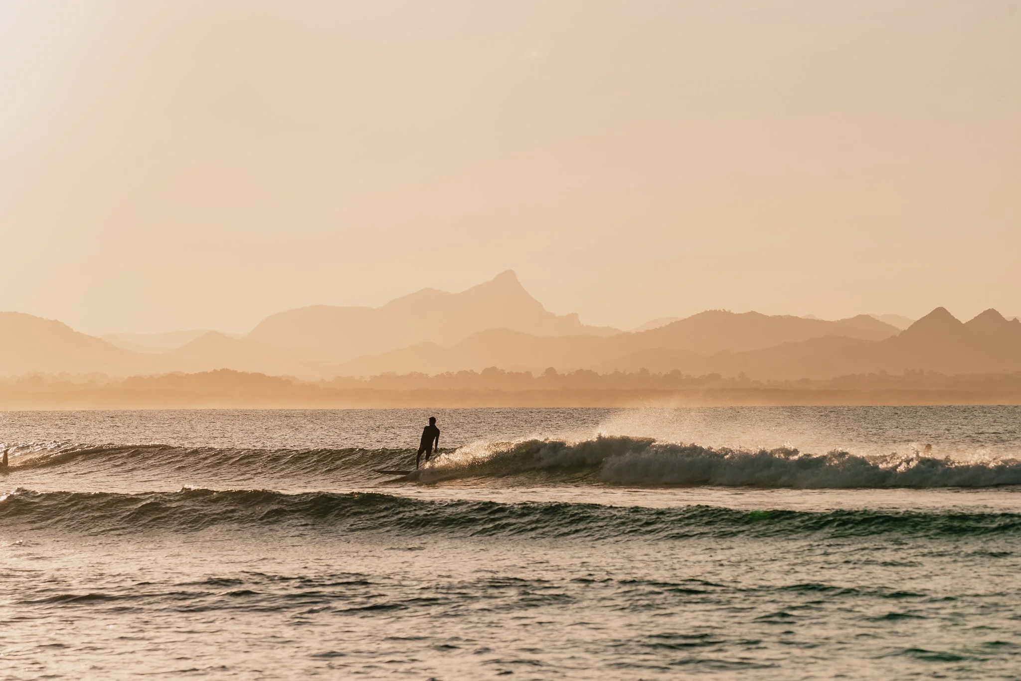A person surfing on a wave in the ocean during sunset with mountains in the background.
