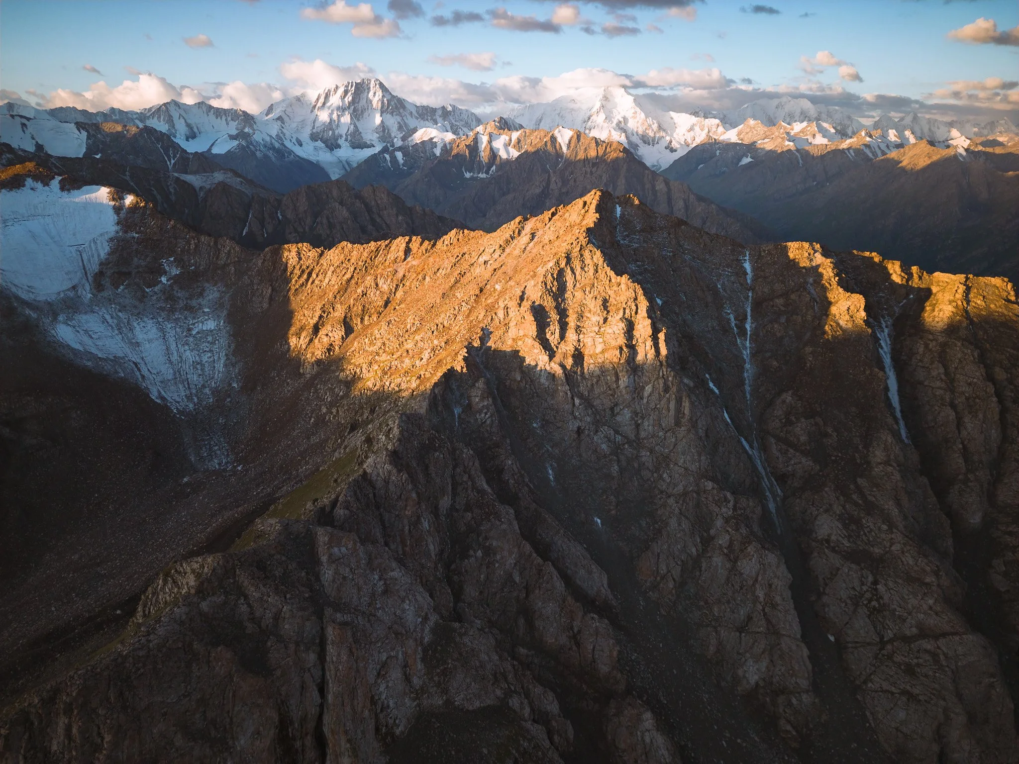 Photograph of rugged mountain landscape with snow-capped peaks in the background, and rocky, steep slopes in the foreground illuminated by golden sunlight, with a few waterfalls cascading down the cliffs.