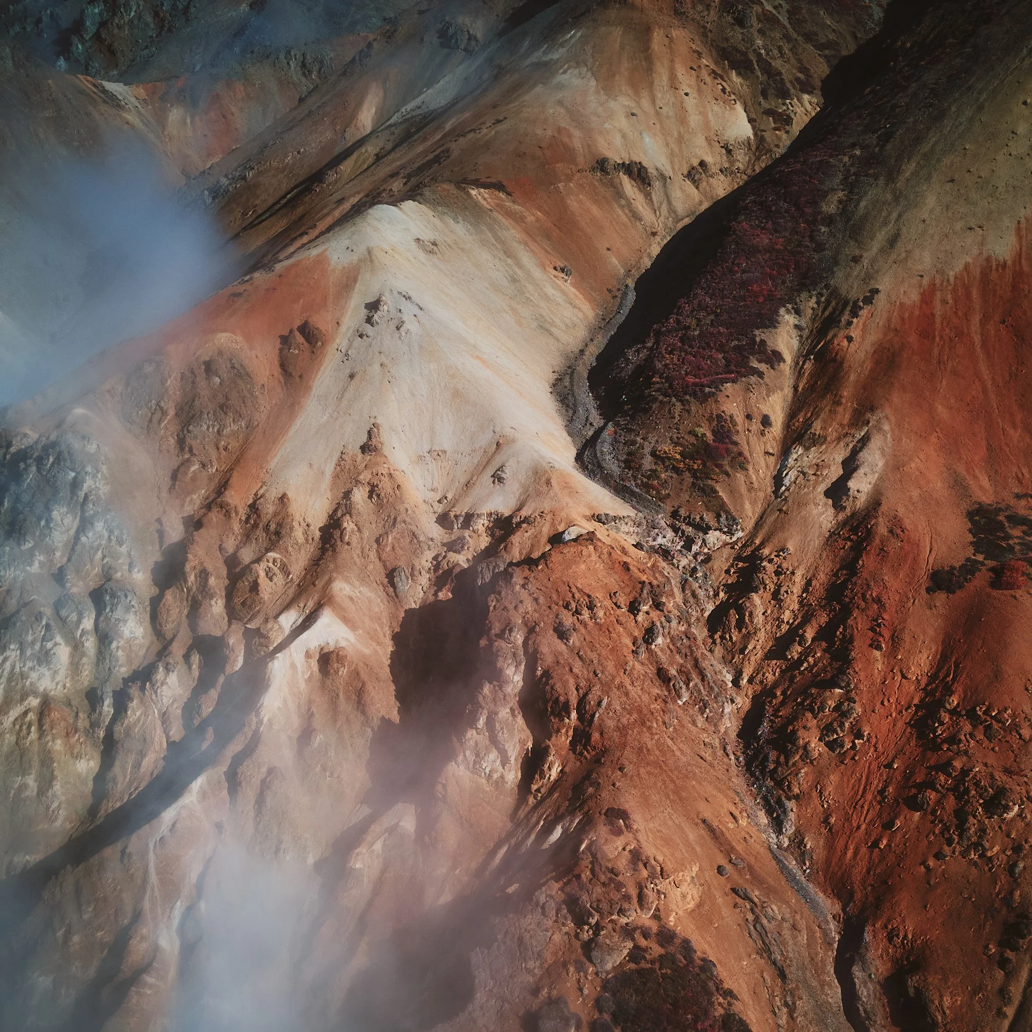 Aerial view of colorful volcanic landscape with steep slopes, layered rock formations, and patches of vegetation, partly obscured by mist.