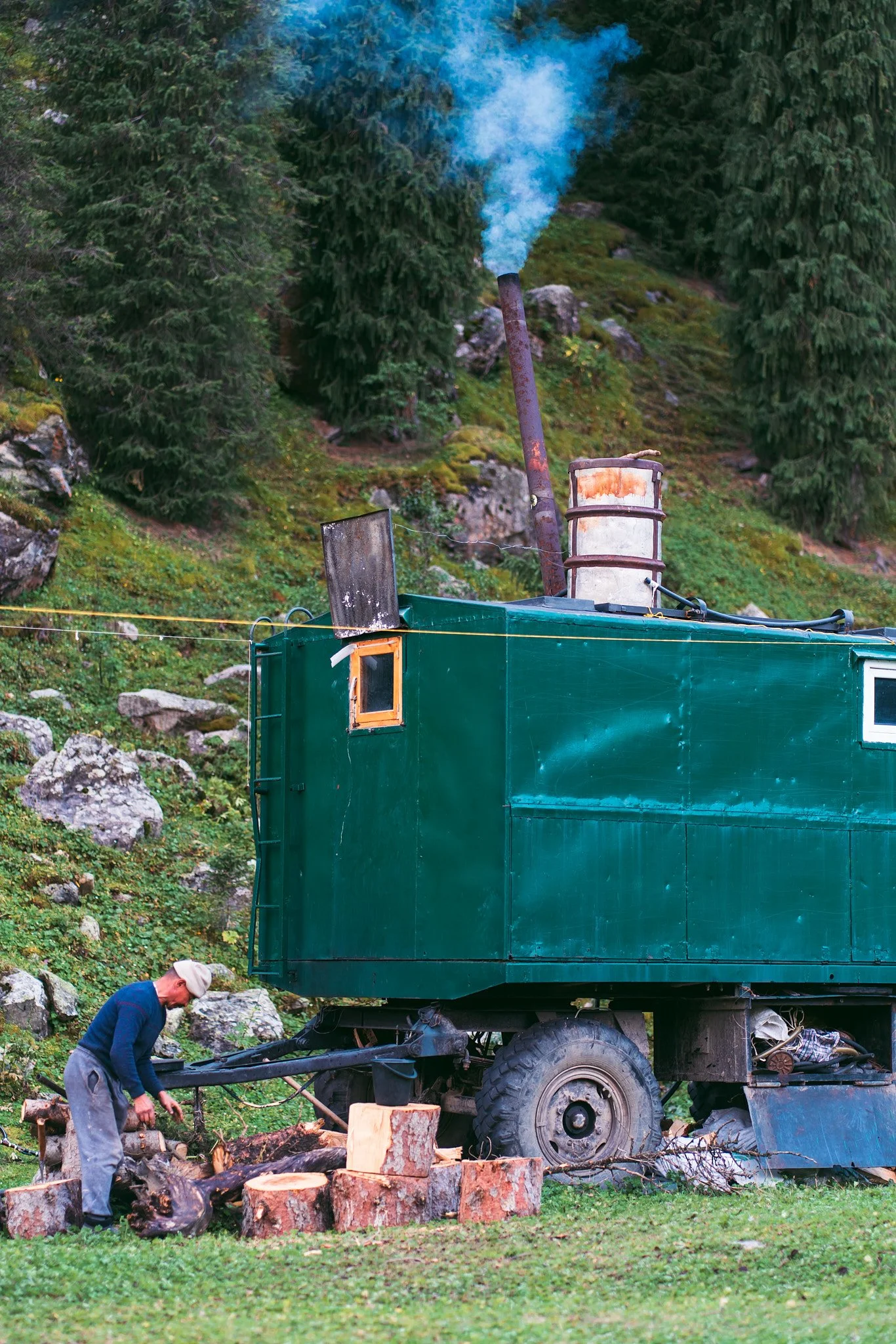 A person chopping wood next to a green mobile home parked on grass in a wooded area.