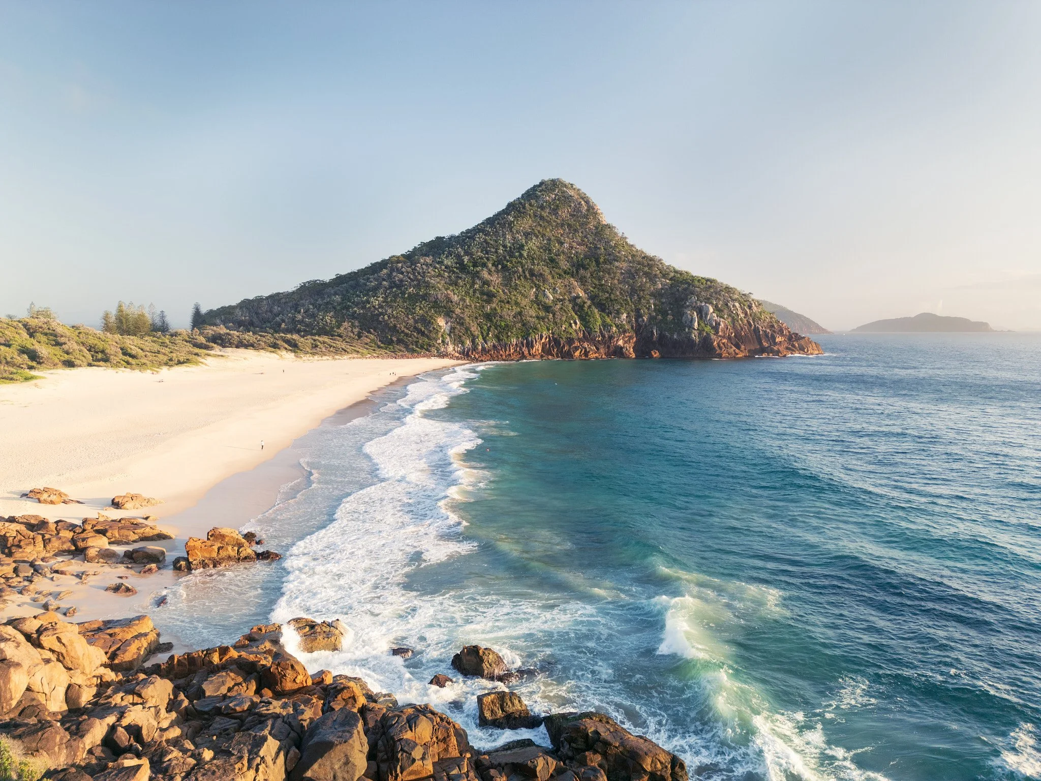A scenic view of a beach with white sand, rocks in the foreground, and a large green hill in the background. The ocean waves are gently crashing onto the shore, and the sky is clear with some light clouds.