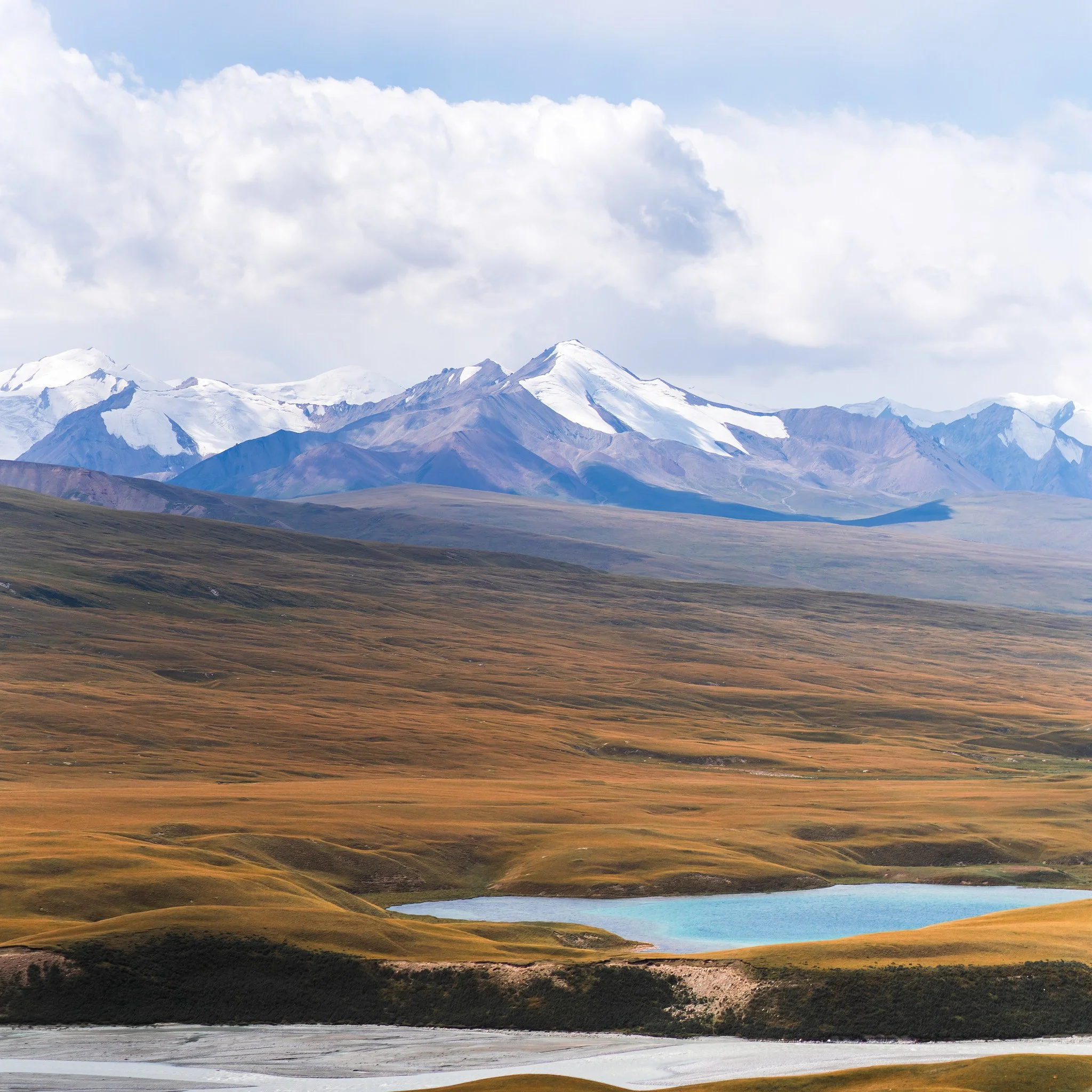 Scenic view of mountains with snow-capped peaks, rolling grassy hills, a small blue lake, and a partly cloudy sky