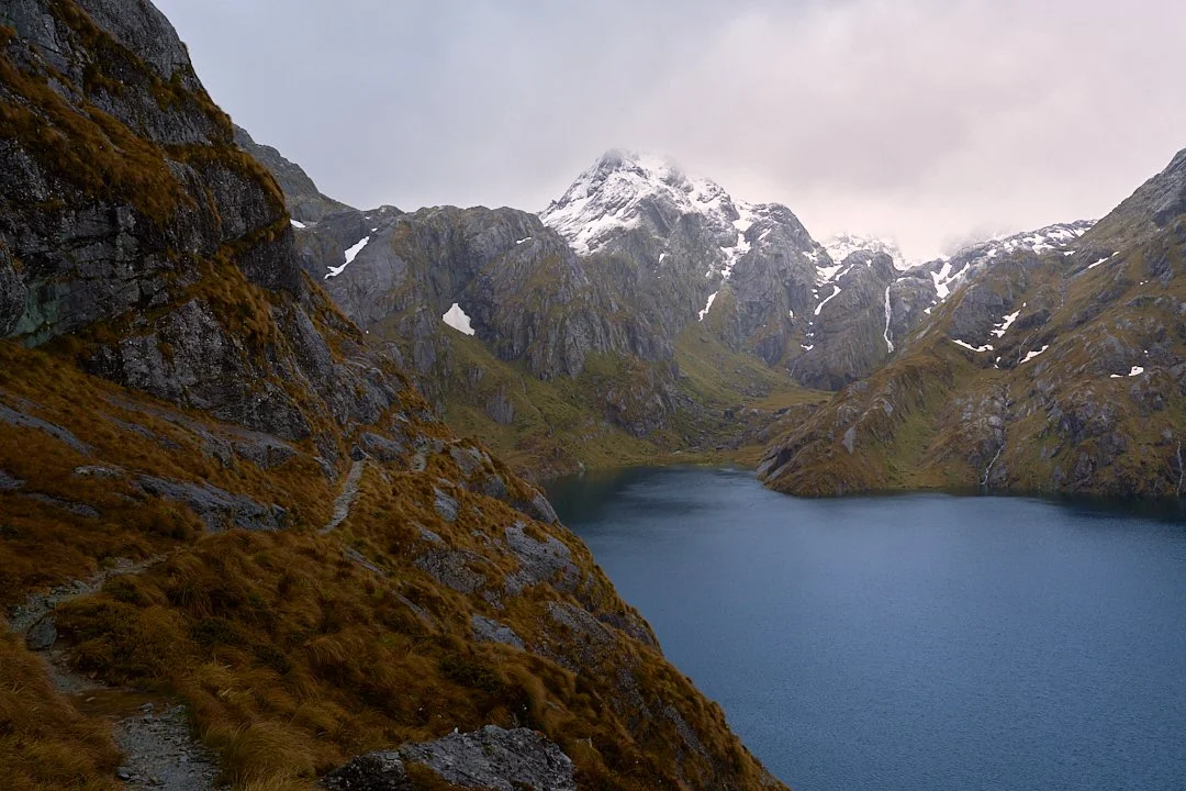 A mountain landscape featuring a large lake surrounded by steep, rocky hills and snow-capped peaks in the background.