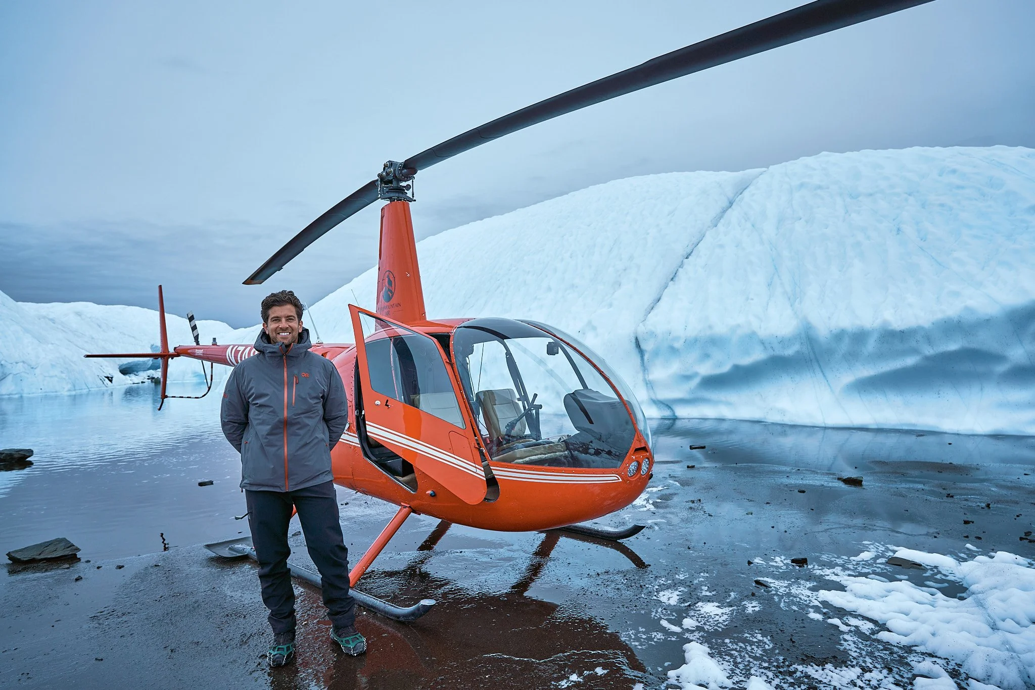 A man in outdoor gear standing next to an orange helicopter on a snowy and icy landscape.