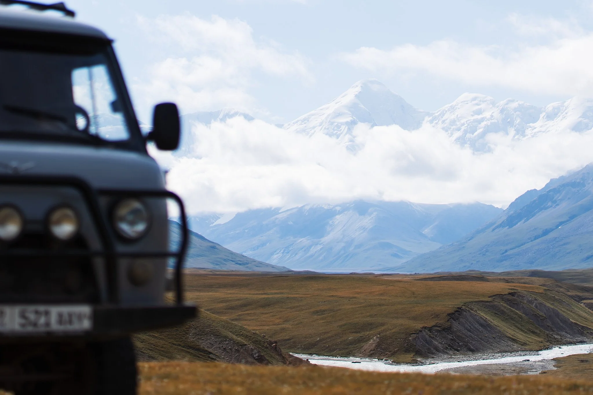 Part of a dark gray off-road truck on the left, with a scenic landscape of mountains, clouds, and a river in the background.