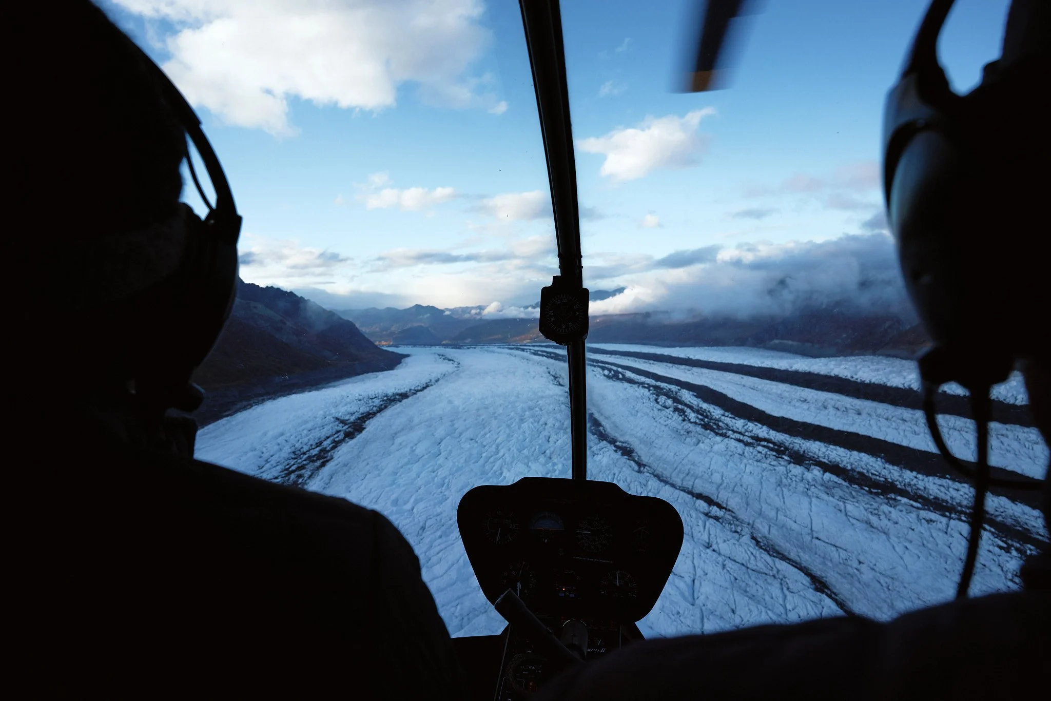 Photo of a helicopter cockpit showing pilots in silhouette flying over snow-covered mountains and a river valley.