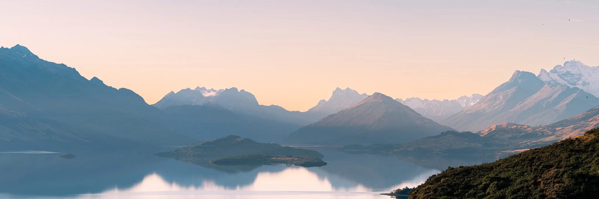 A serene landscape of a mountain range reflected in a calm lake at sunset or sunrise, with a pastel-colored sky and snow-capped peaks in the distance.