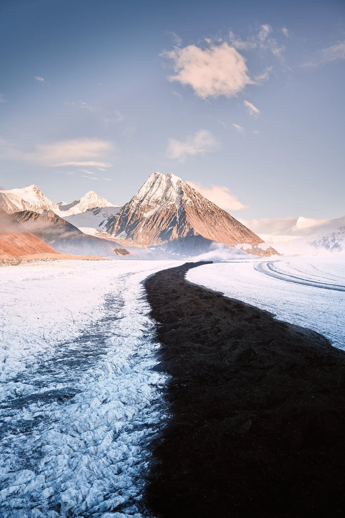 Snow-covered landscape with a dark dirt path leading towards a snowy mountain range in the distance.
