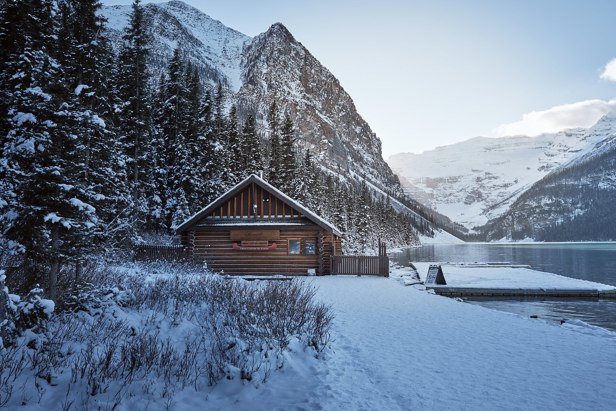 A snow-covered landscape features a small wooden cabin beside a lake, surrounded by snowy mountains and dense pine trees under a clear blue sky.