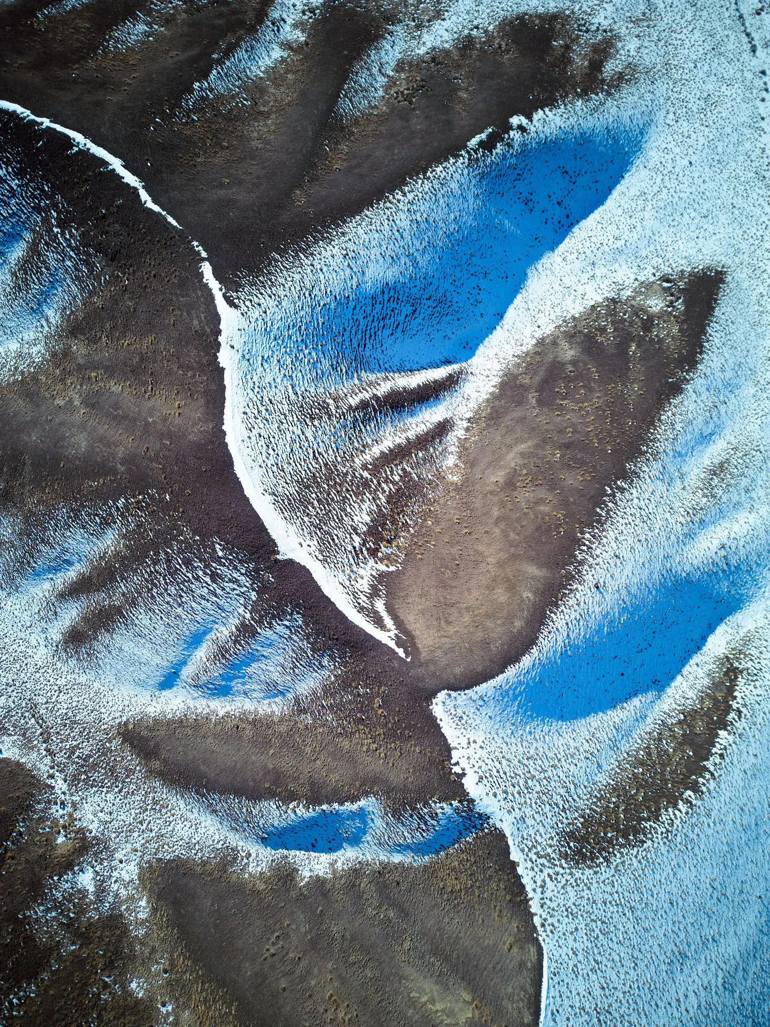 An aerial view of a landscape with dark soil, patches of snow or ice, and areas of blue water, creating abstract patterns.