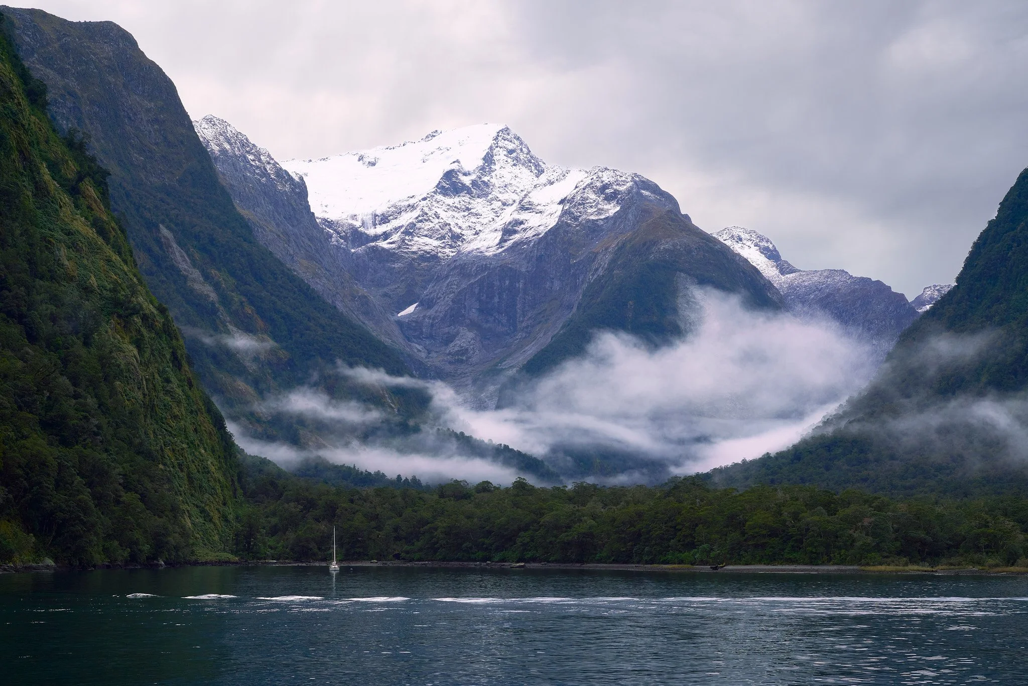 Scenic view of a fjord with calm water, lush green hills, and tall snow-capped mountains in the background, with mist and clouds in the sky.