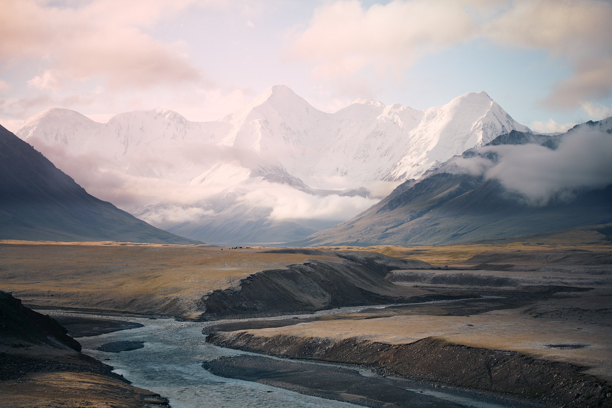 Snow-capped mountains in the background with clouds, a valley with a river flowing through, grassy plains, and some grazing animals.
