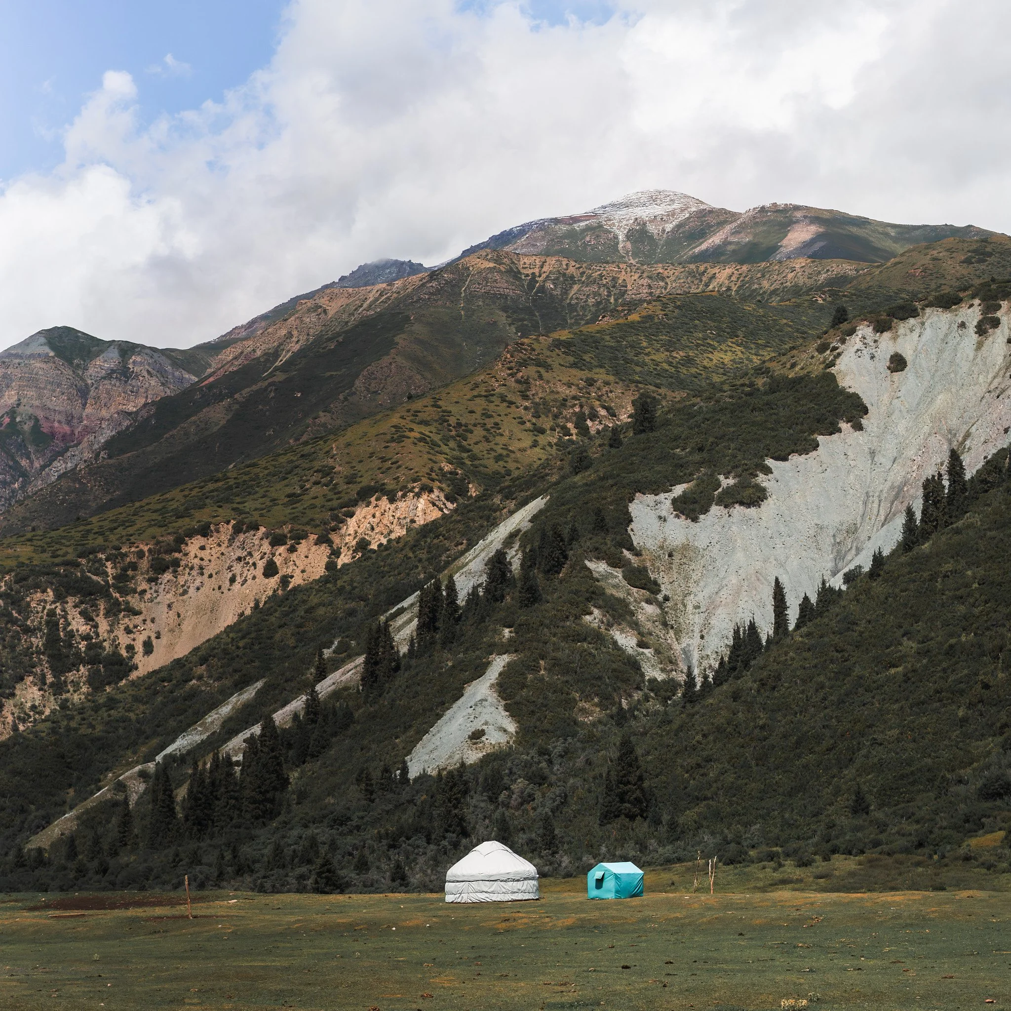 A mountainous landscape with two tents, a white yurt and a blue tent, set on a grassy field in front of steep, green, rocky mountains with slopes covered with trees and shrubs.