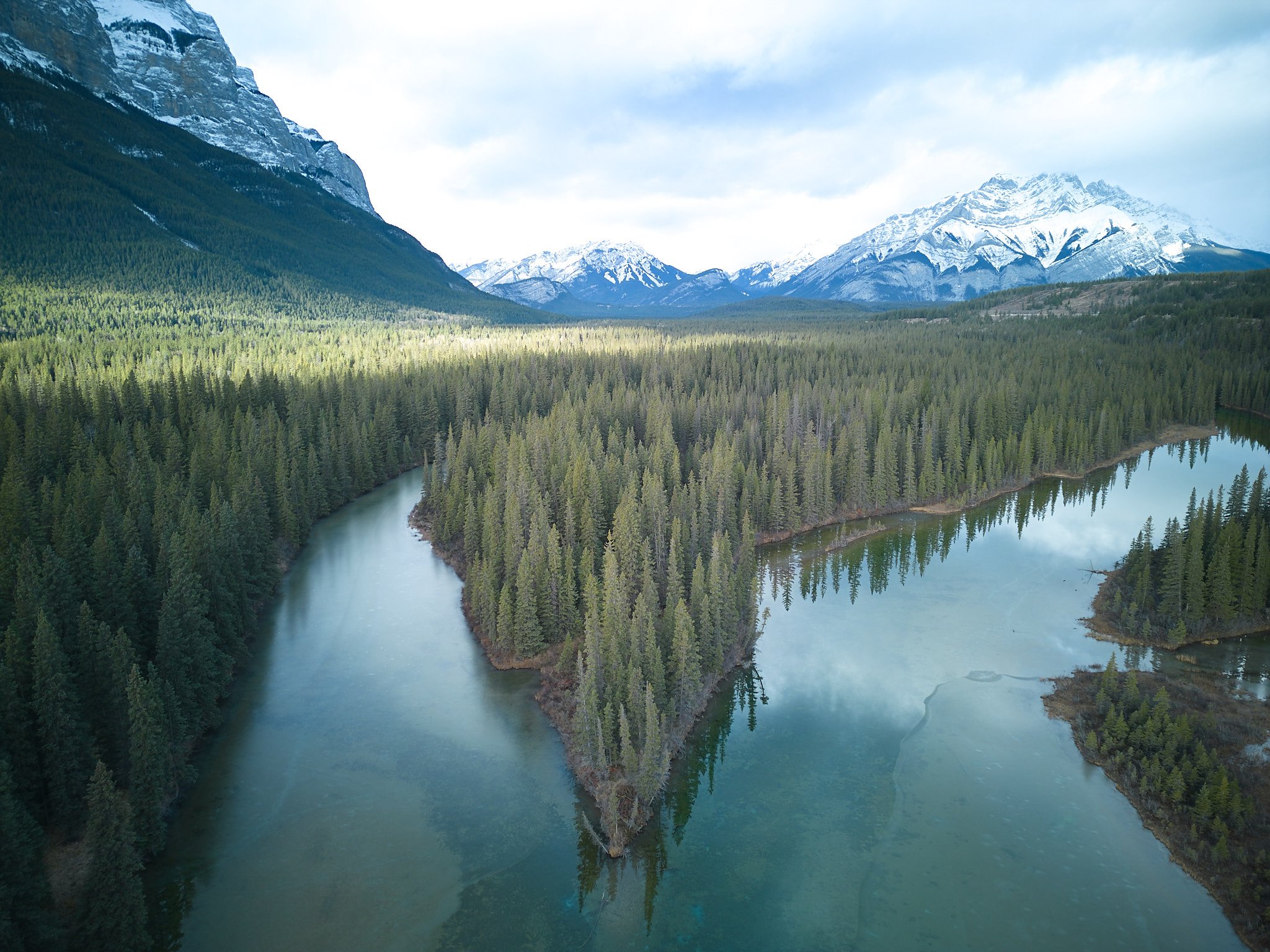Aerial view of a river winding through a dense forest with snow-capped mountains in the background.