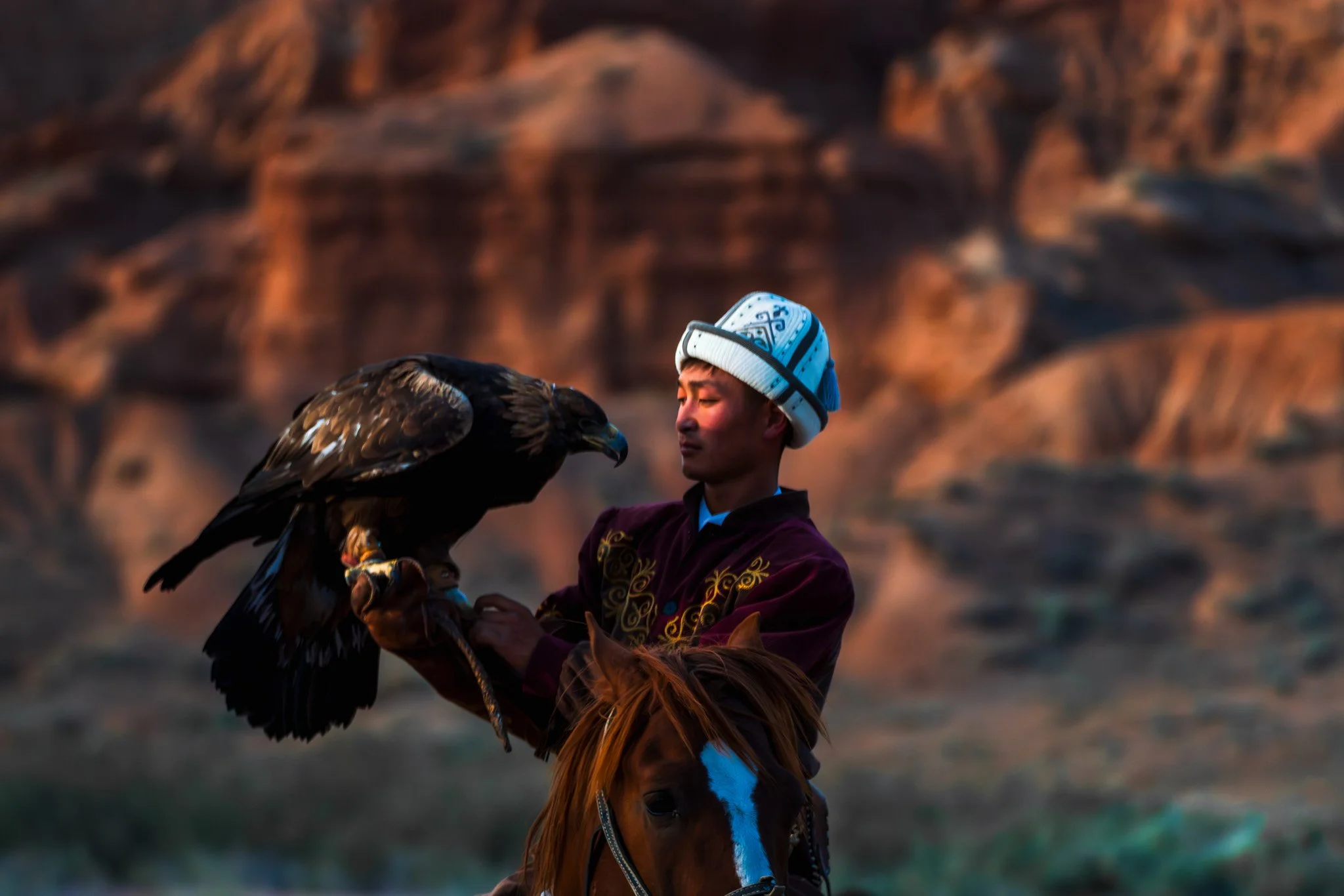 A man in traditional attire holding a large bird of prey on horseback against a mountainous landscape at sunset.
