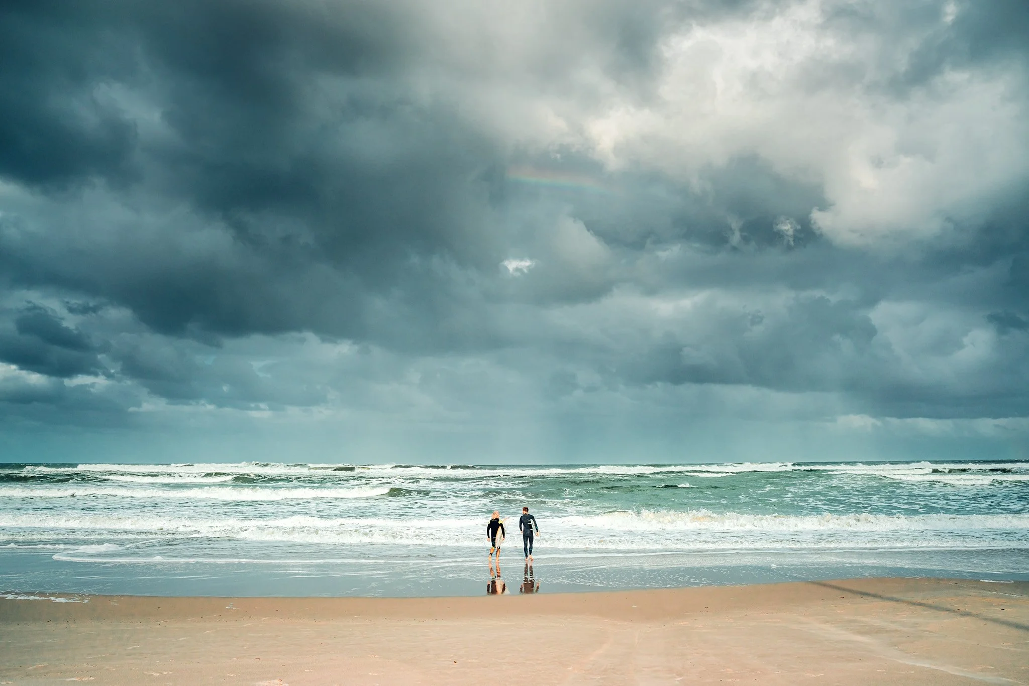 Two people walking on the sandy beach towards the ocean with dark stormy clouds overhead and a faint rainbow in the sky.