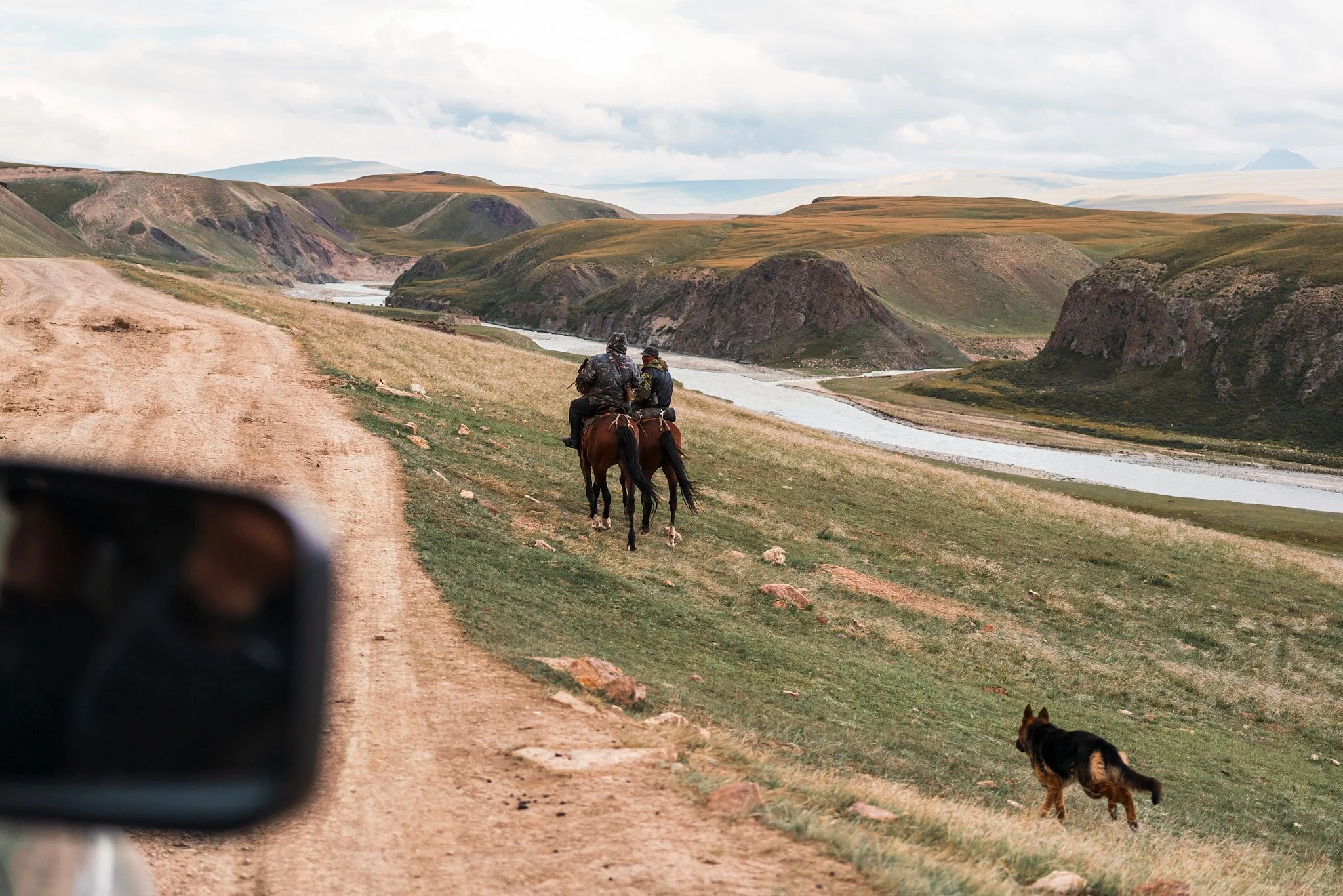Two people riding horses along a dirt road in a rural, mountainous landscape with a river flowing through the valley, a dog walking on the grass nearby, and cloudy sky overhead.