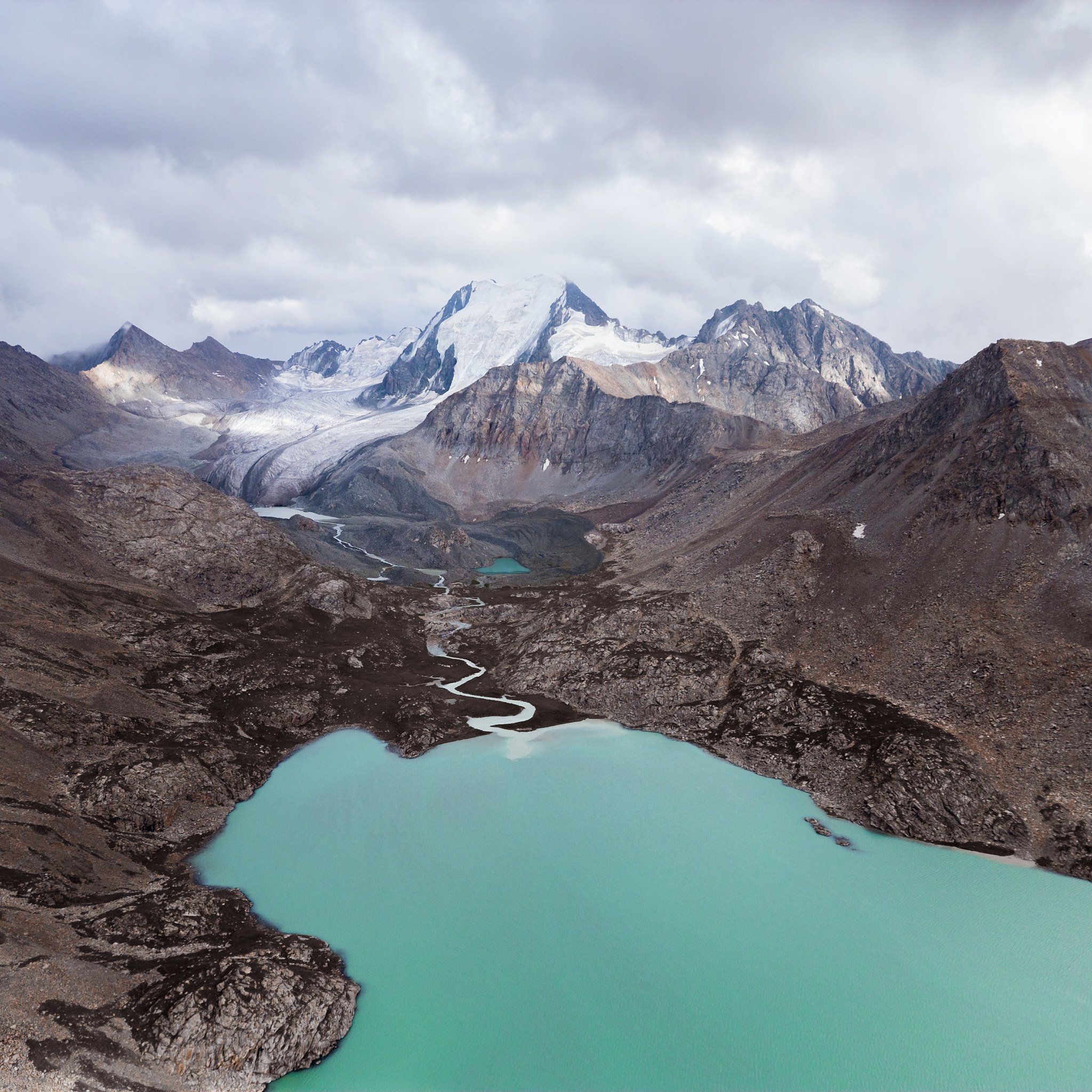 A mountain landscape with snow-capped peaks, rocky slopes, and two turquoise lakes connected by a winding stream under a cloudy sky.