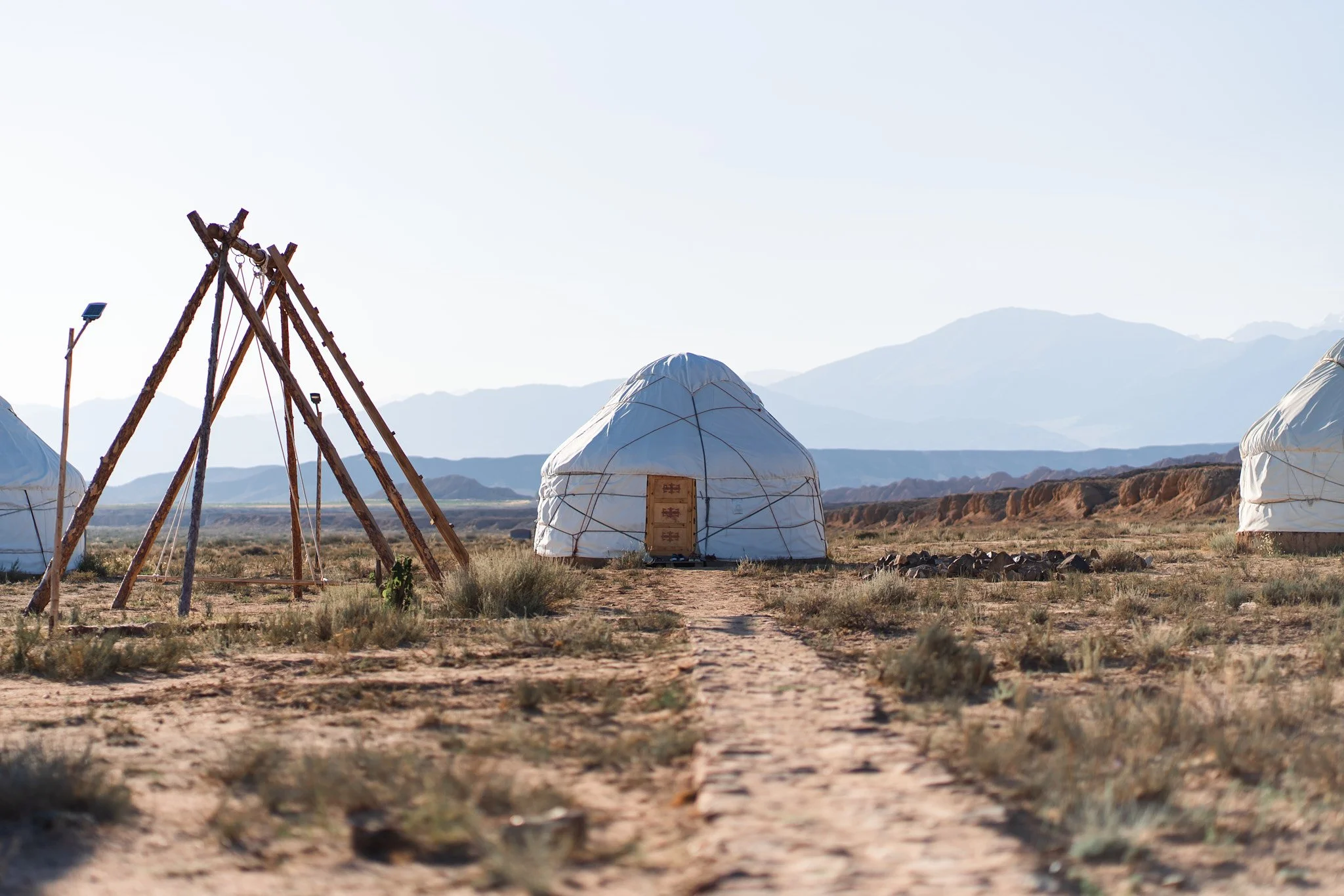 A desert landscape with three white yurts, wooden teepee structure, distant mountains, and a dirt path.