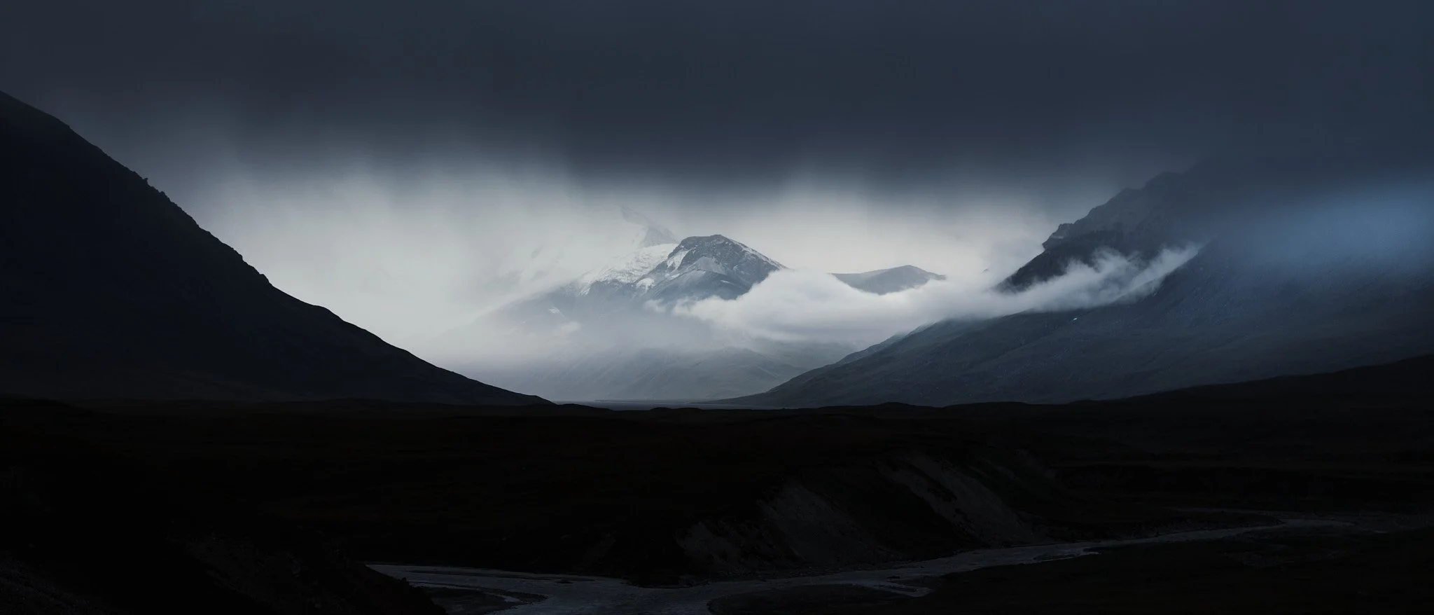 Dark mountain landscape with snow-capped peaks and foggy clouds.
