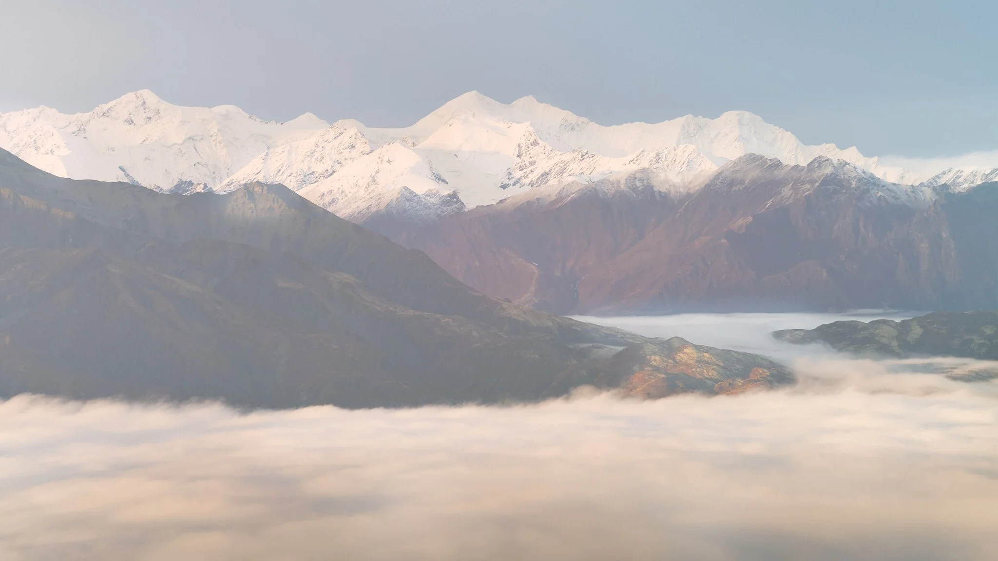 Snow-capped mountains with rugged dark hills in the foreground and clouds below.