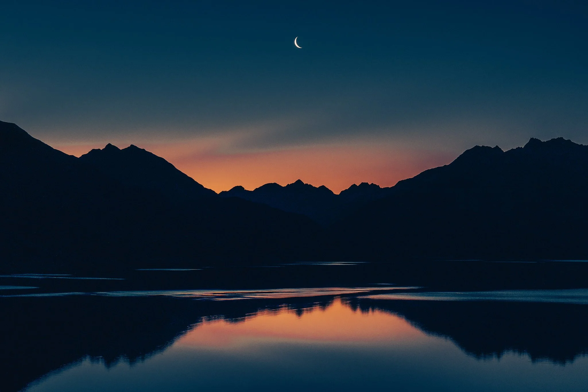 Nighttime mountain landscape with a crescent moon in the sky and a calm lake reflecting the mountains and sky.