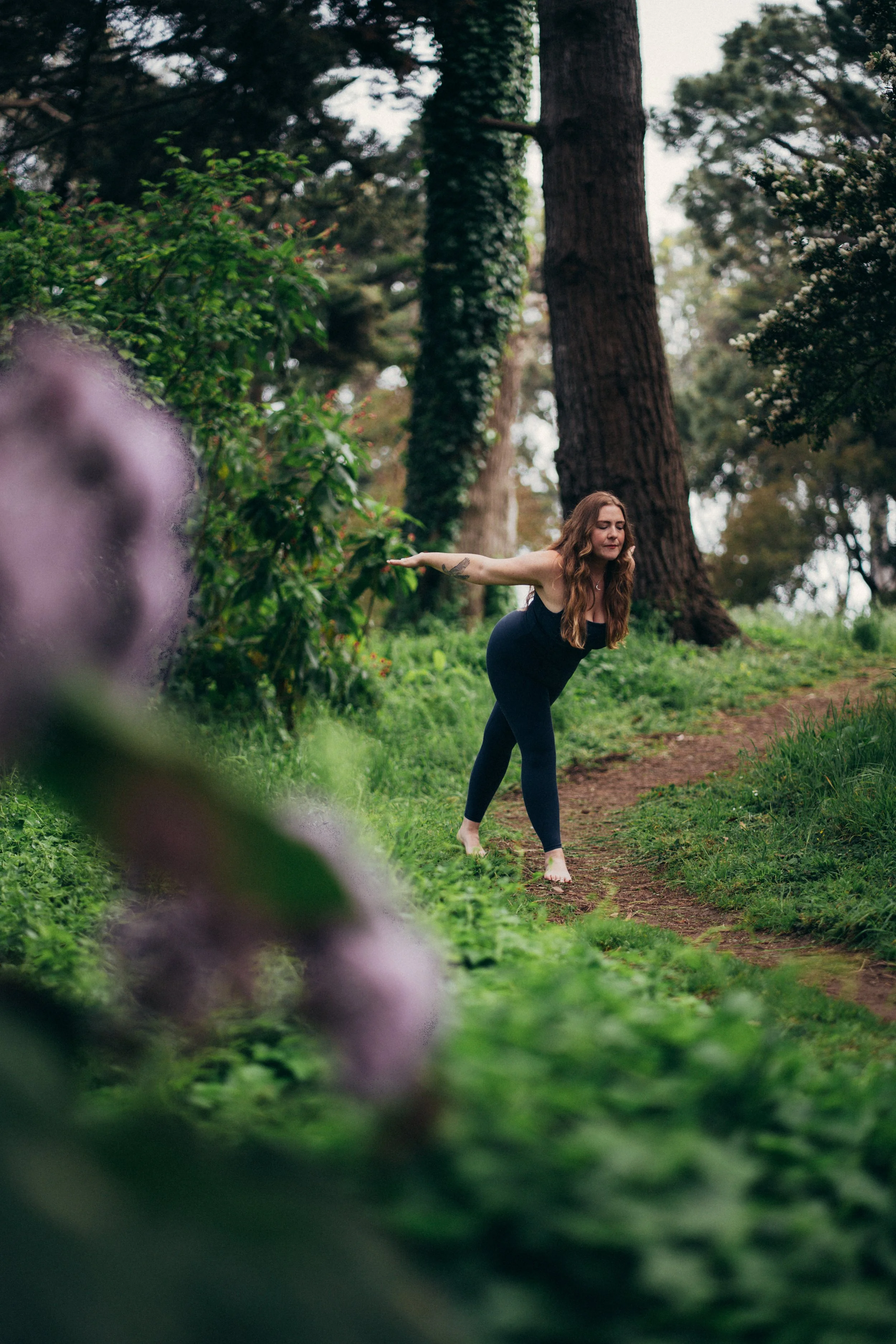 A woman doing yoga outdoors in a lush, green forest on a dirt path.