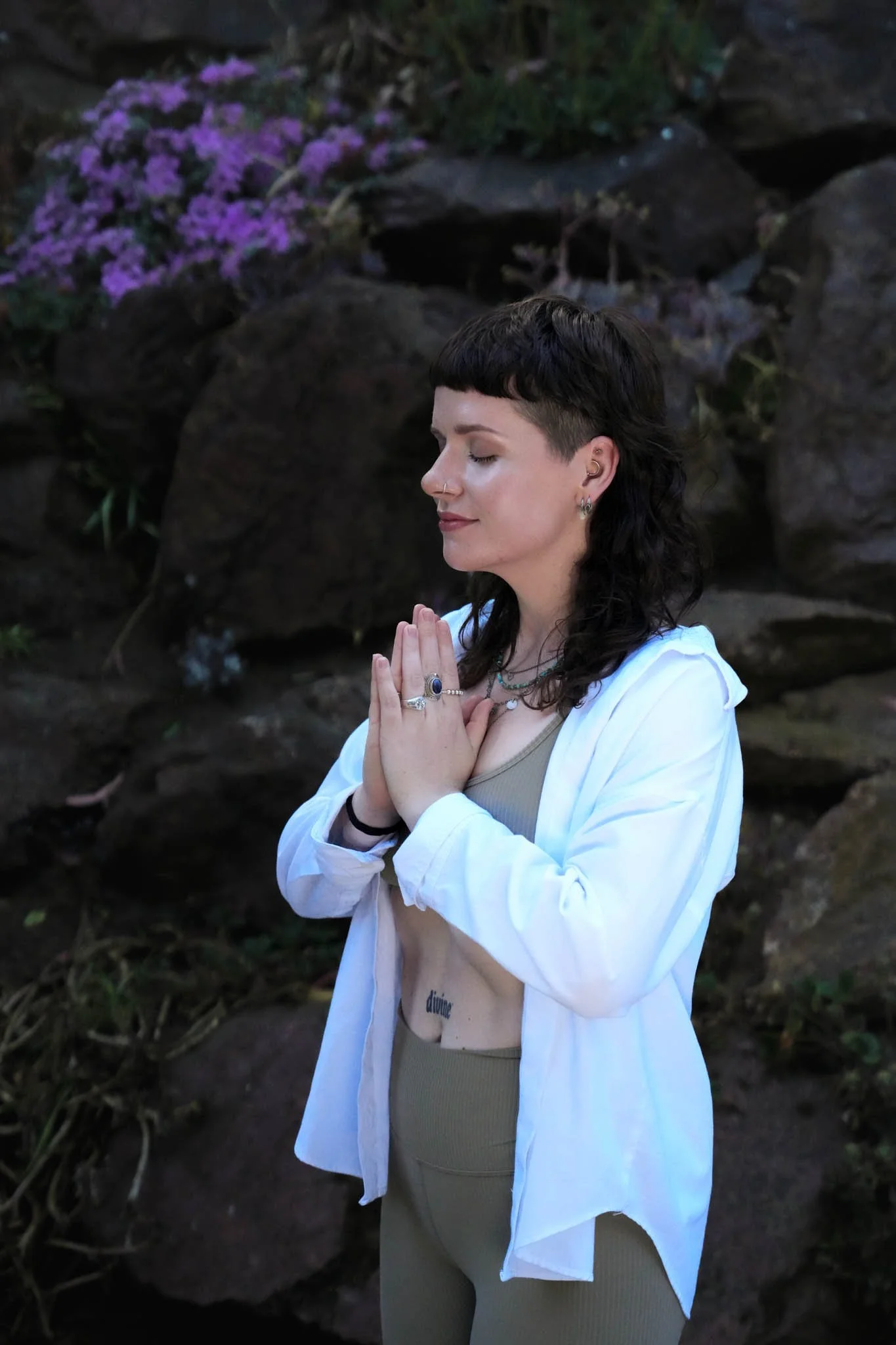 A woman with short dark hair, wearing a white open shirt over beige athletic wear, stands outdoors with her eyes closed and hands pressed together in a prayer position. She is surrounded by rocks and pink flowers.