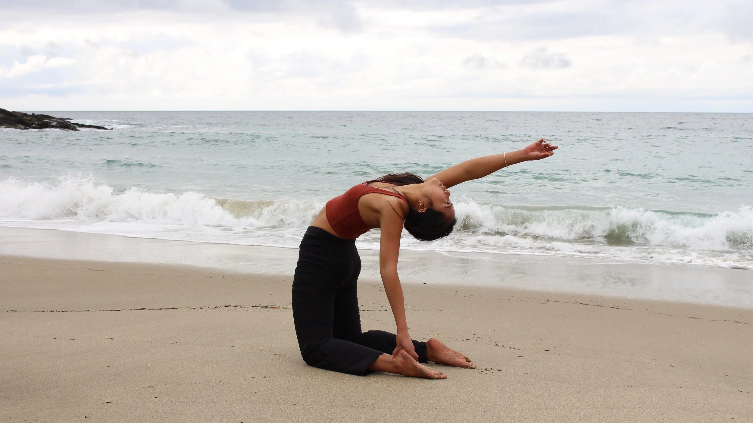 Woman practicing yoga on the beach, in a kneeling pose with her head and arm extended backward.