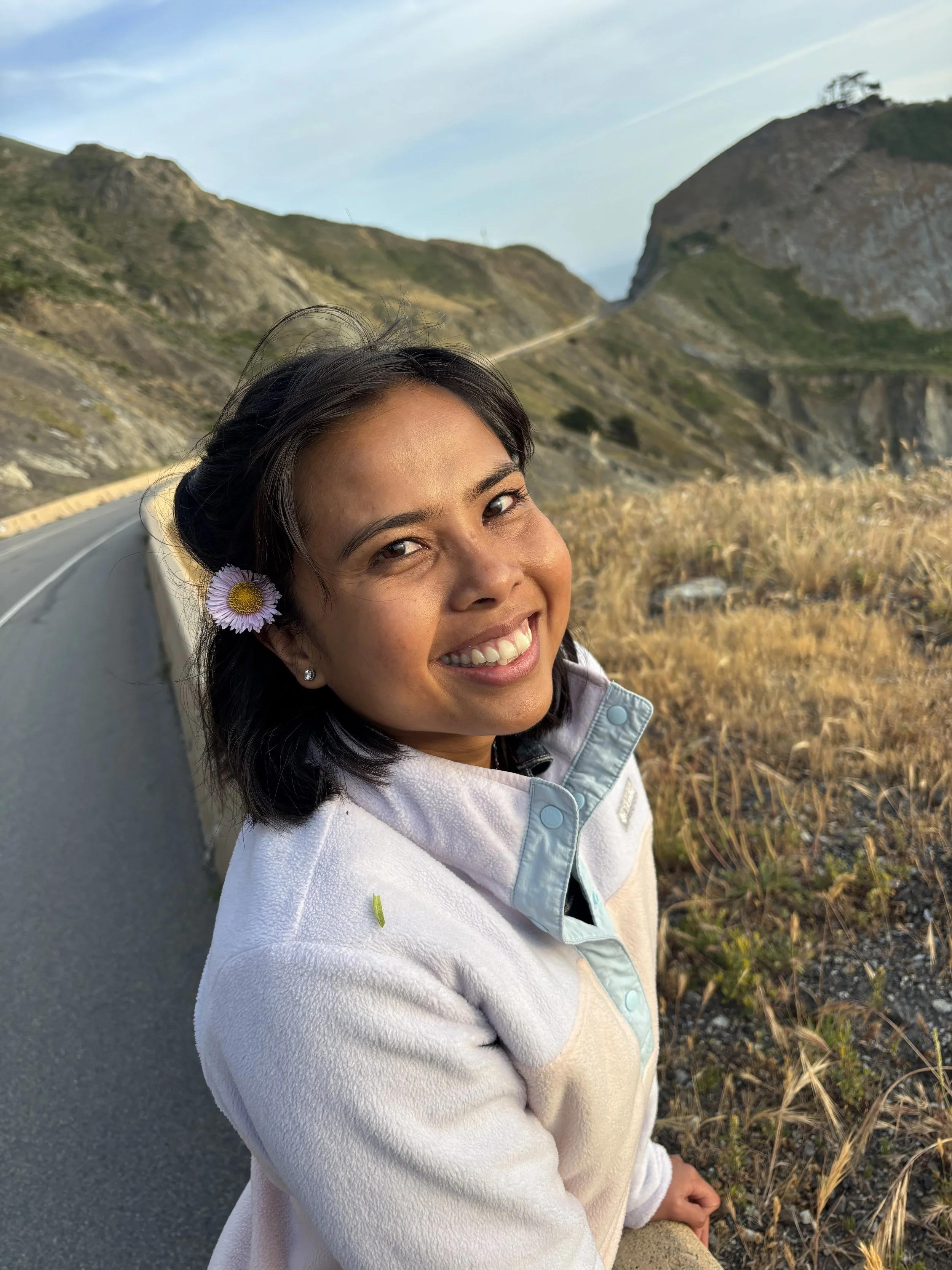 A woman smiling with a purple daisy in her hair, standing by a winding mountain road with hills and dry grass in the background during daytime.