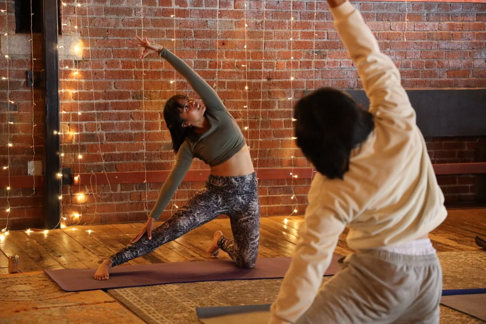 A woman doing yoga in a brick-walled studio with string lights, with another person in the foreground reaching upward.