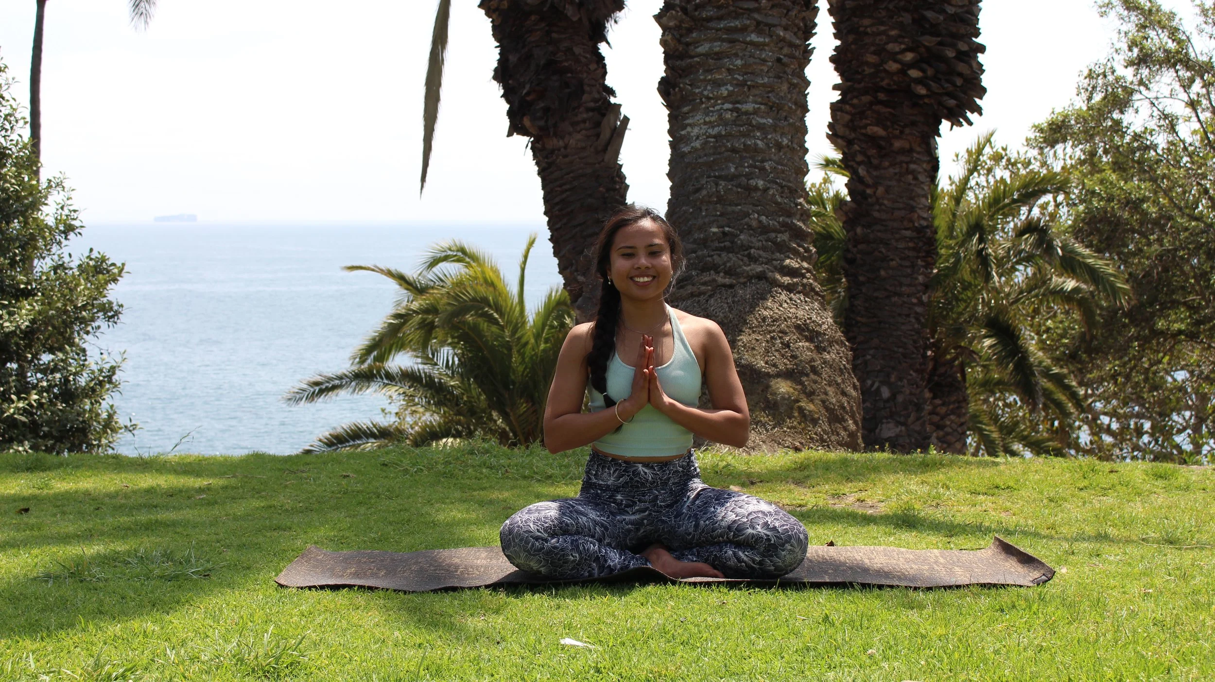A young woman practicing yoga outdoors on a grassy area near palm trees and a view of the ocean, sitting cross-legged with hands in prayer position and smiling.