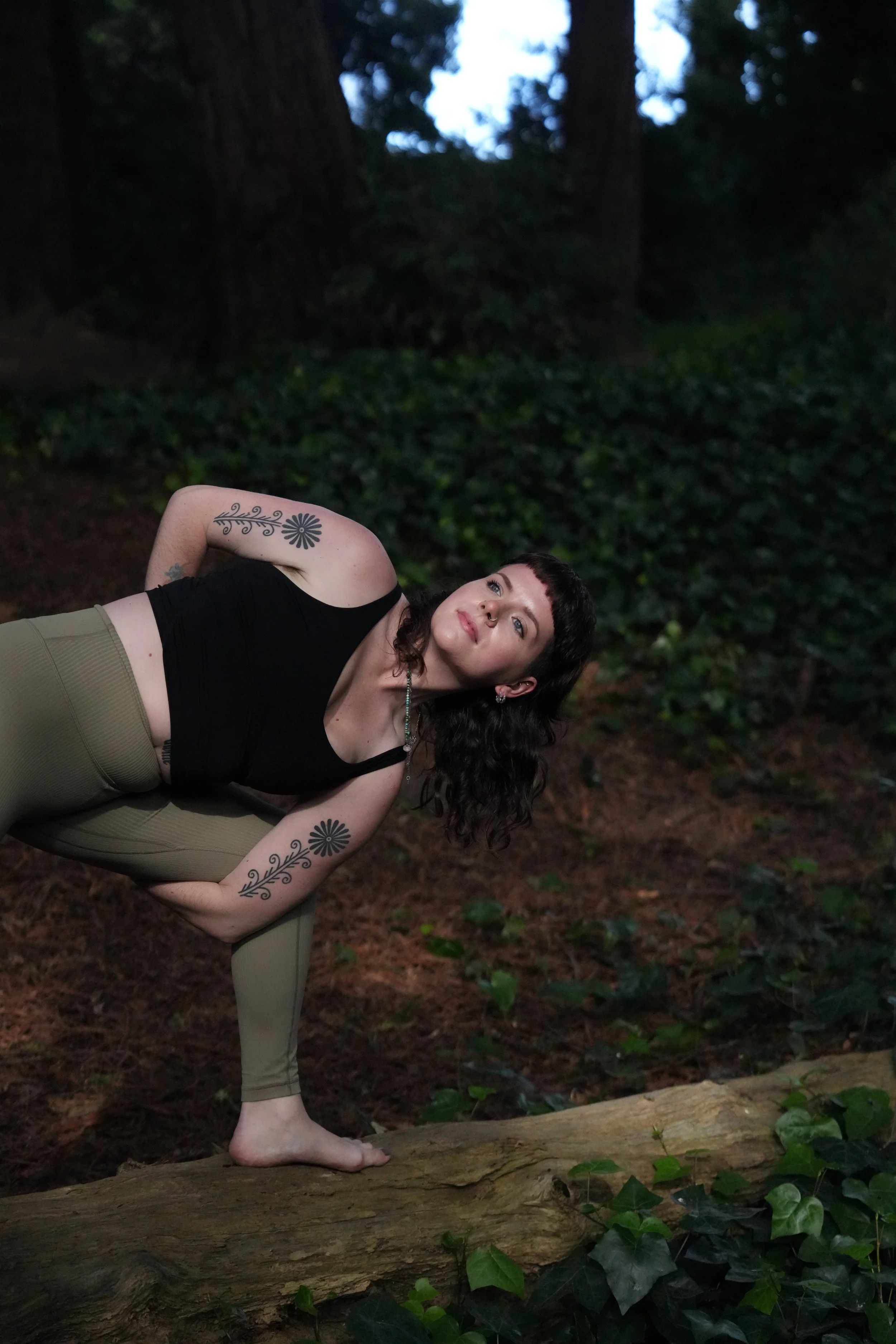 A woman with dark, curly hair is practicing yoga outdoors on a fallen log, surrounded by dense greenery and trees during dusk.