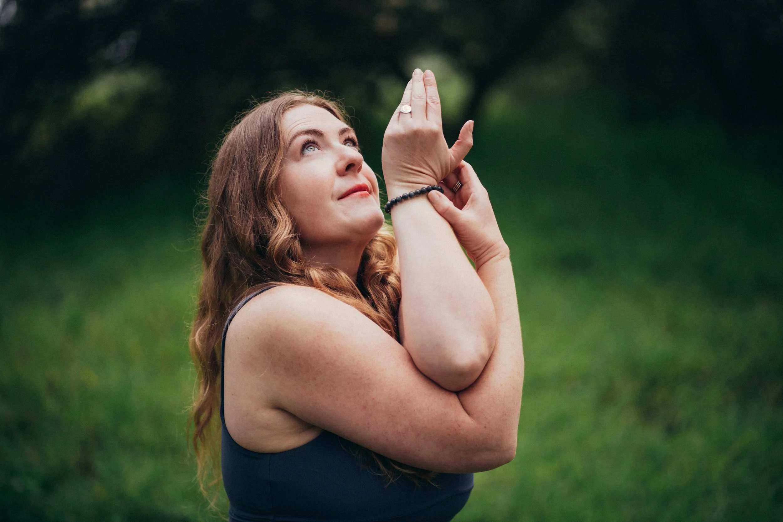 A woman with long, wavy red hair practicing yoga outdoors on a green grassy field, with her hands pressed together in a prayer position and looking upwards.